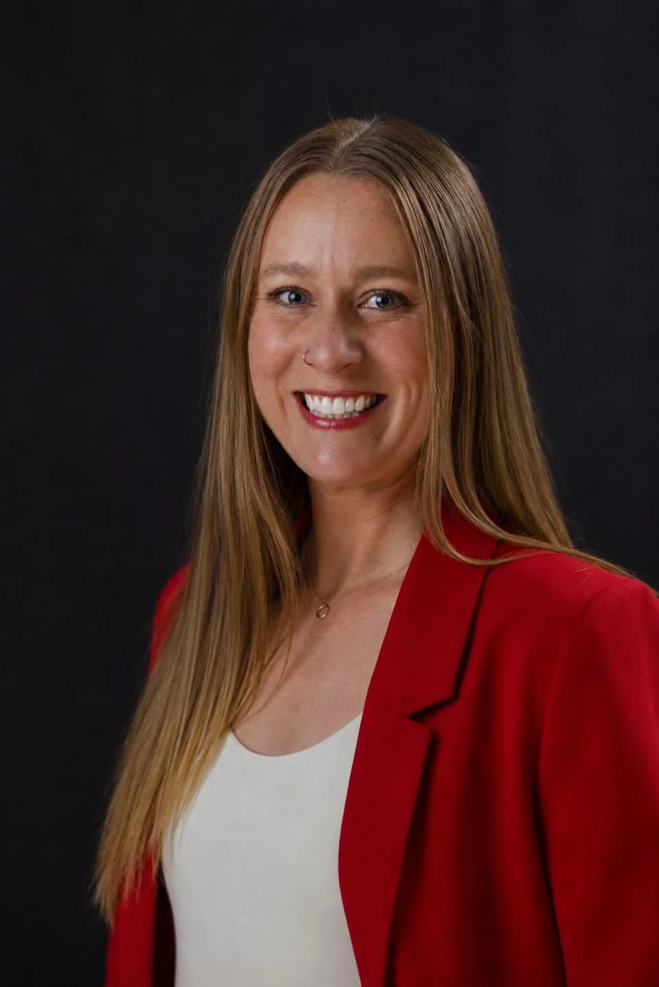 Woman with long red hair, smiling, wearing a red blazer and a white top, against a dark background.