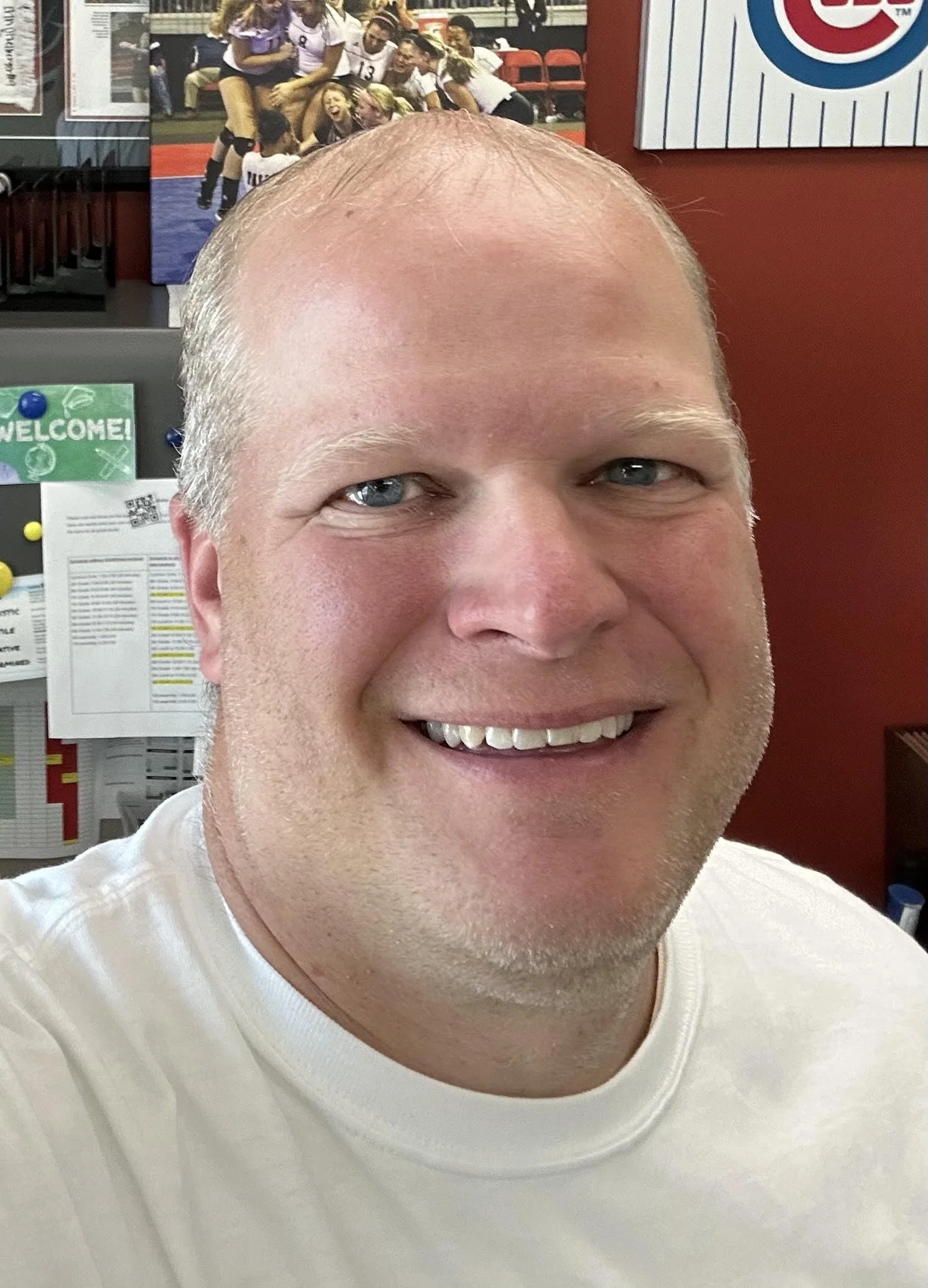 Close-up of a smiling man with a bald head and blue eyes, wearing a white shirt, with a sports-themed poster featuring volleyball players in the background.