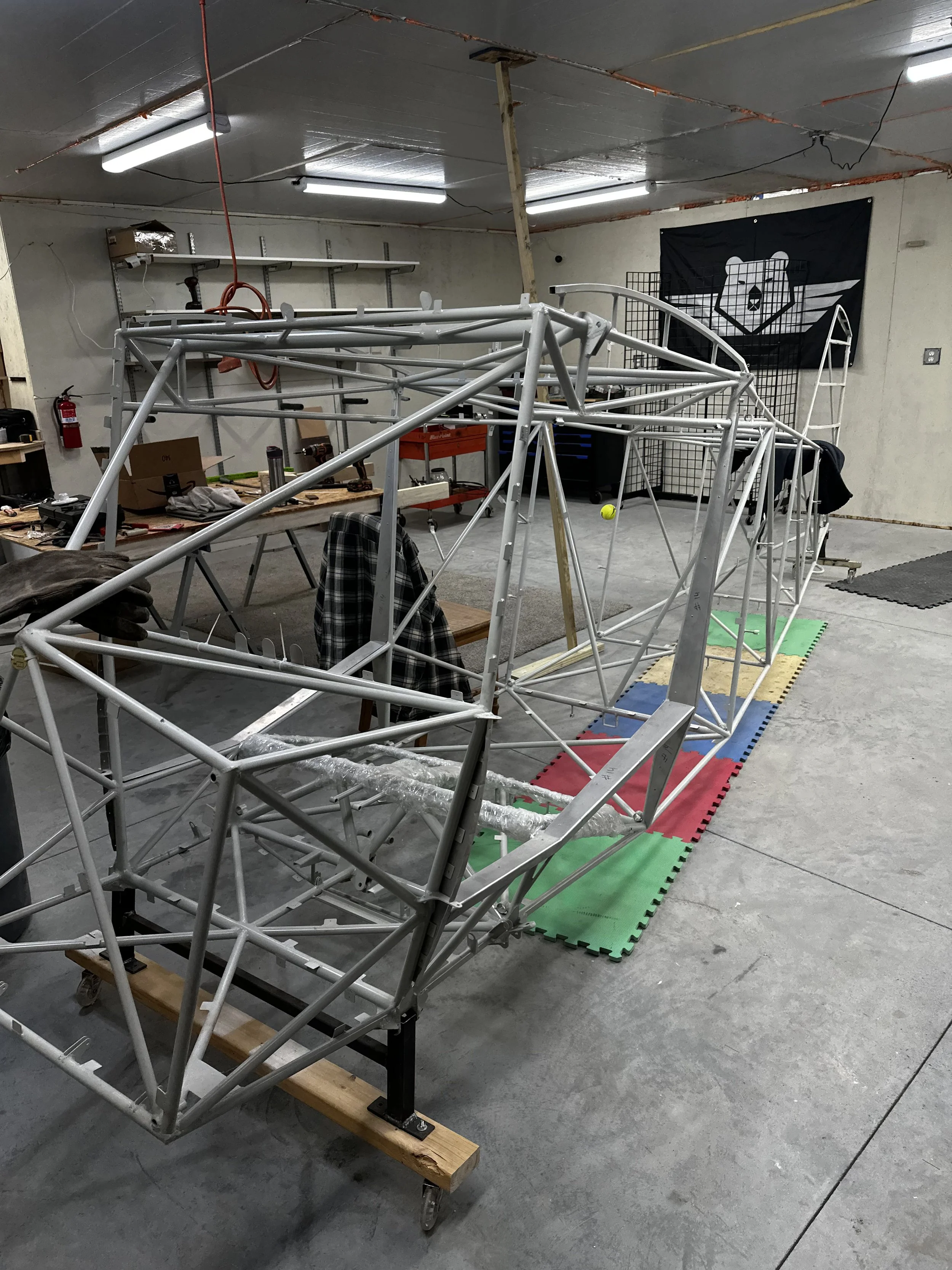 A skeletal frame of a small vehicle or robot is mounted on a wooden platform with caster wheels in a workshop with tools, workbenches, shelves, and a black flag featuring a bear face with wings in the background.