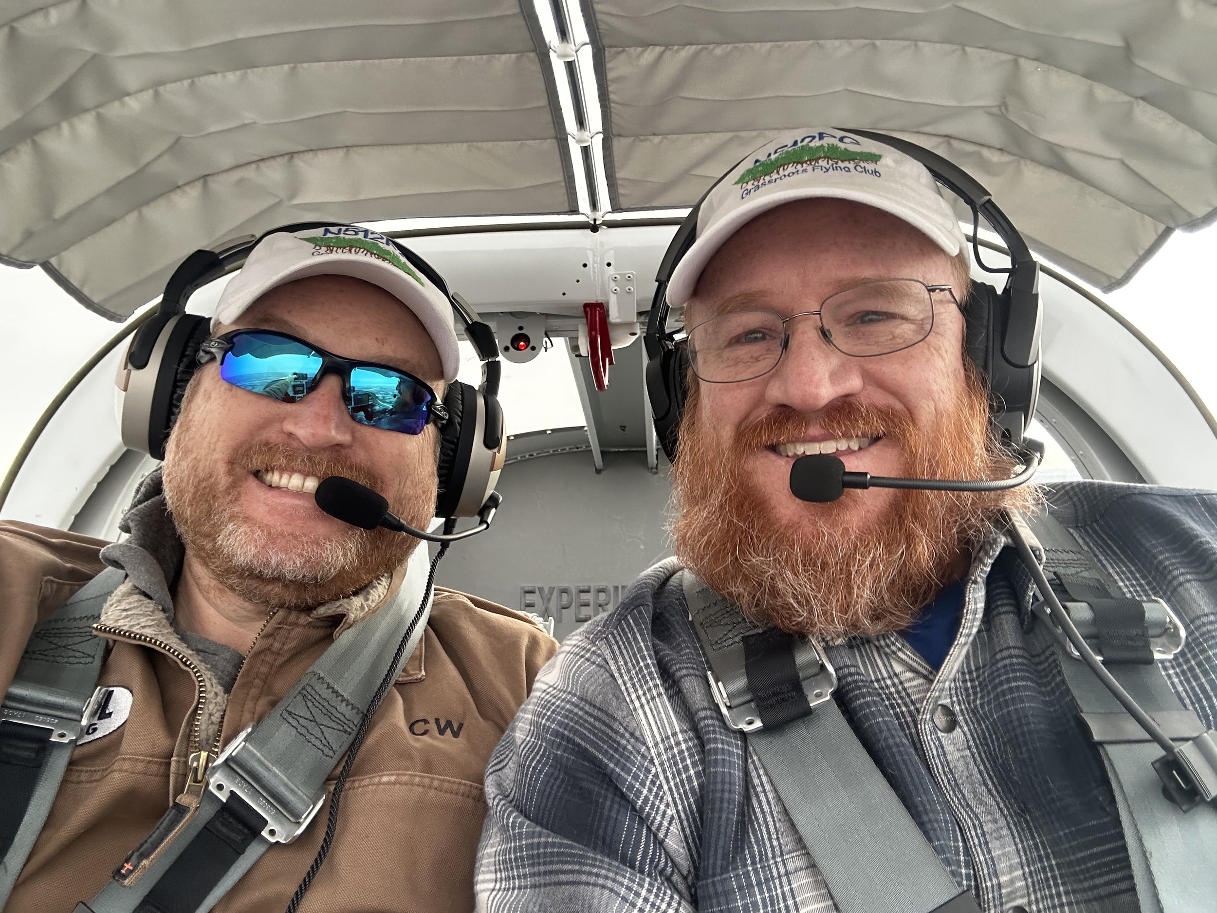 Two men with red hair and beards wearing white caps and headsets, smiling inside an aircraft cockpit, with one wearing glasses and the other sunglasses.
