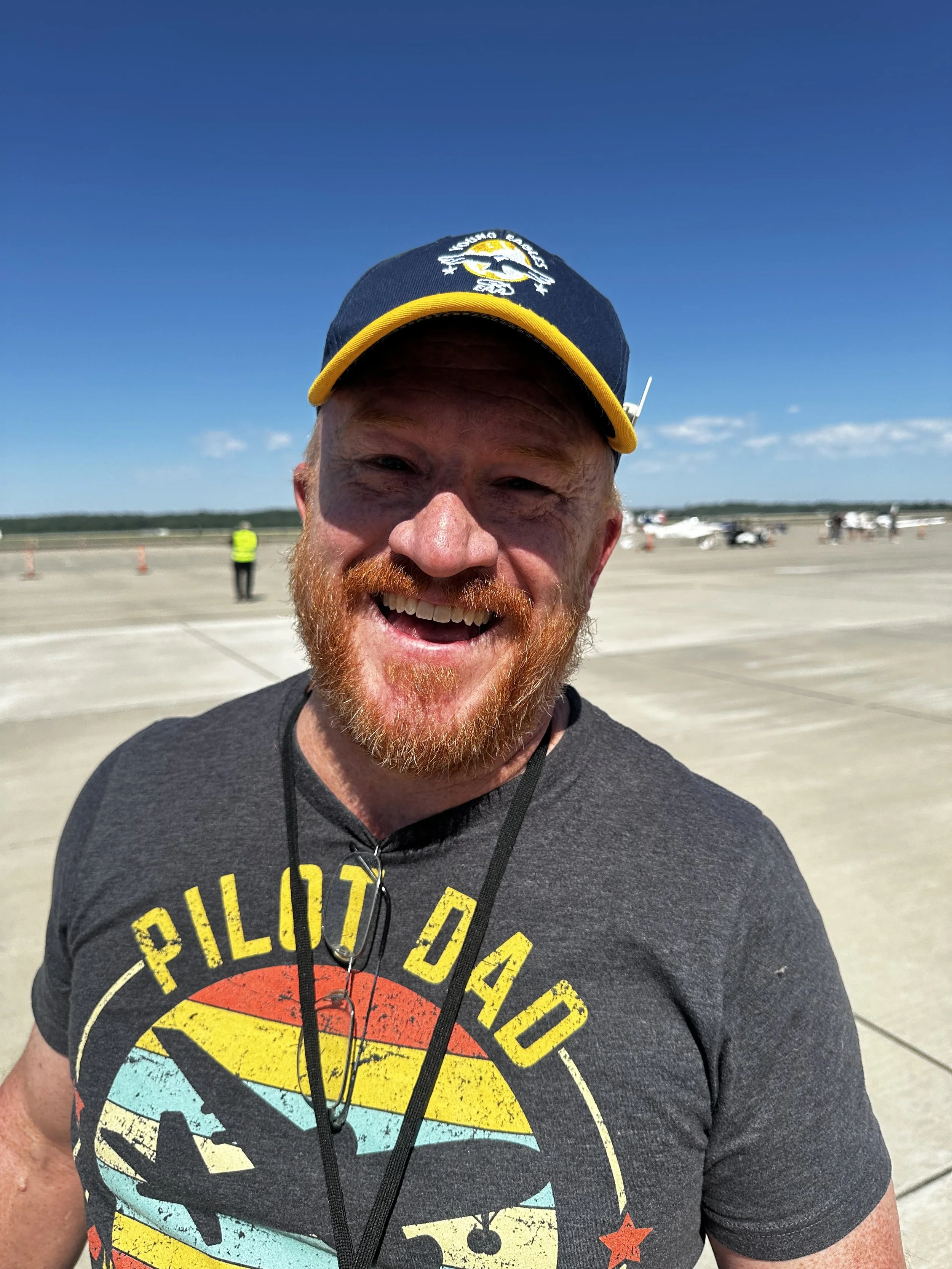 A man with a red beard smiling, wearing a black t-shirt with a colorful graphic, a blue and yellow baseball cap, and earphones hanging around his neck. The background shows a clear blue sky and an open tarmac with some people and small aircraft in the distance.