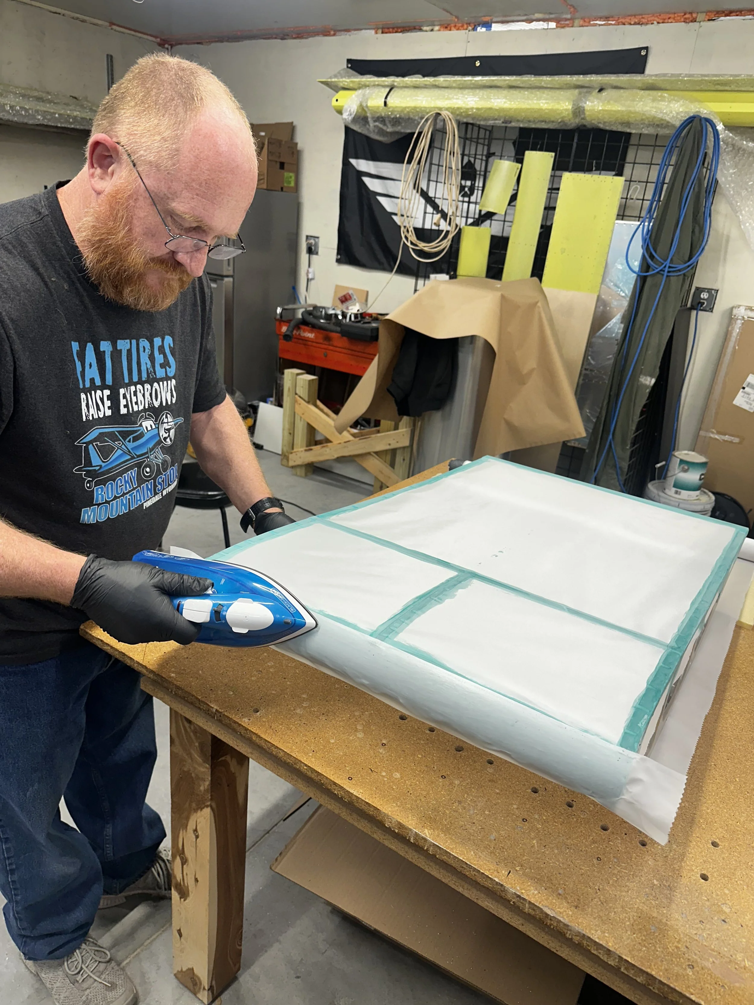 A man with red hair, glasses, and a beard is standing at a workshop table, carefully ironing a piece of fabric or paper that is taped to the table, while wearing black gloves. The workshop has various tools, wires, and materials in the background.