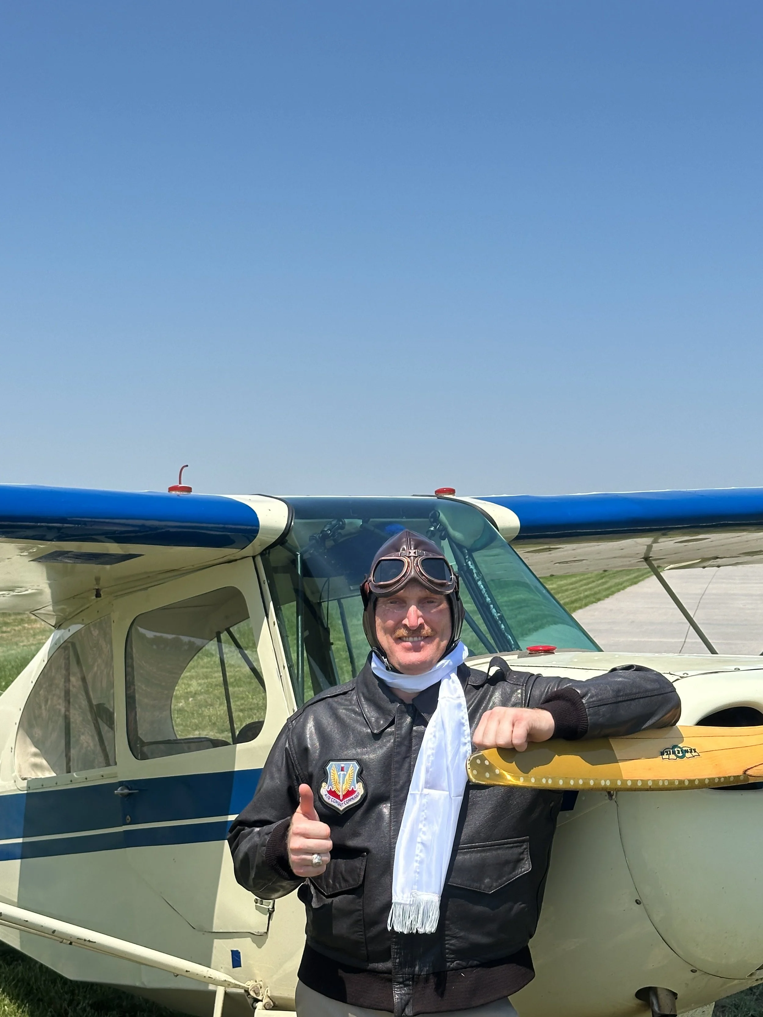 A man wearing vintage pilot goggles and a leather jacket with a patch on it, smiling and giving a thumbs-up, is standing in front of a small light airplane on a sunny day.