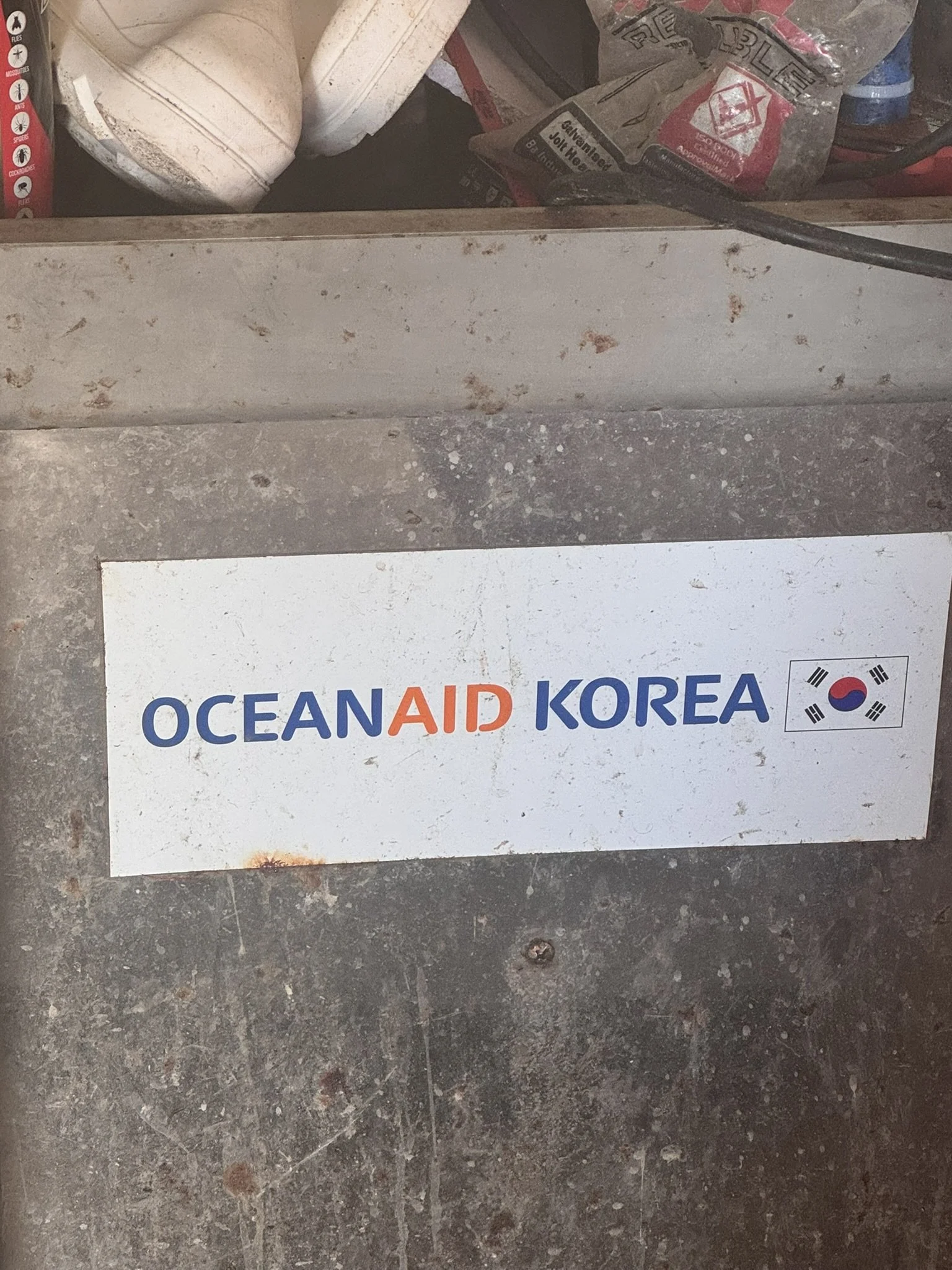 A white sign with the text 'OCEAN AID KOREA' and the South Korean flag symbol on it, placed on a dirty, speckled concrete floor with various construction or cleaning supplies in the background.