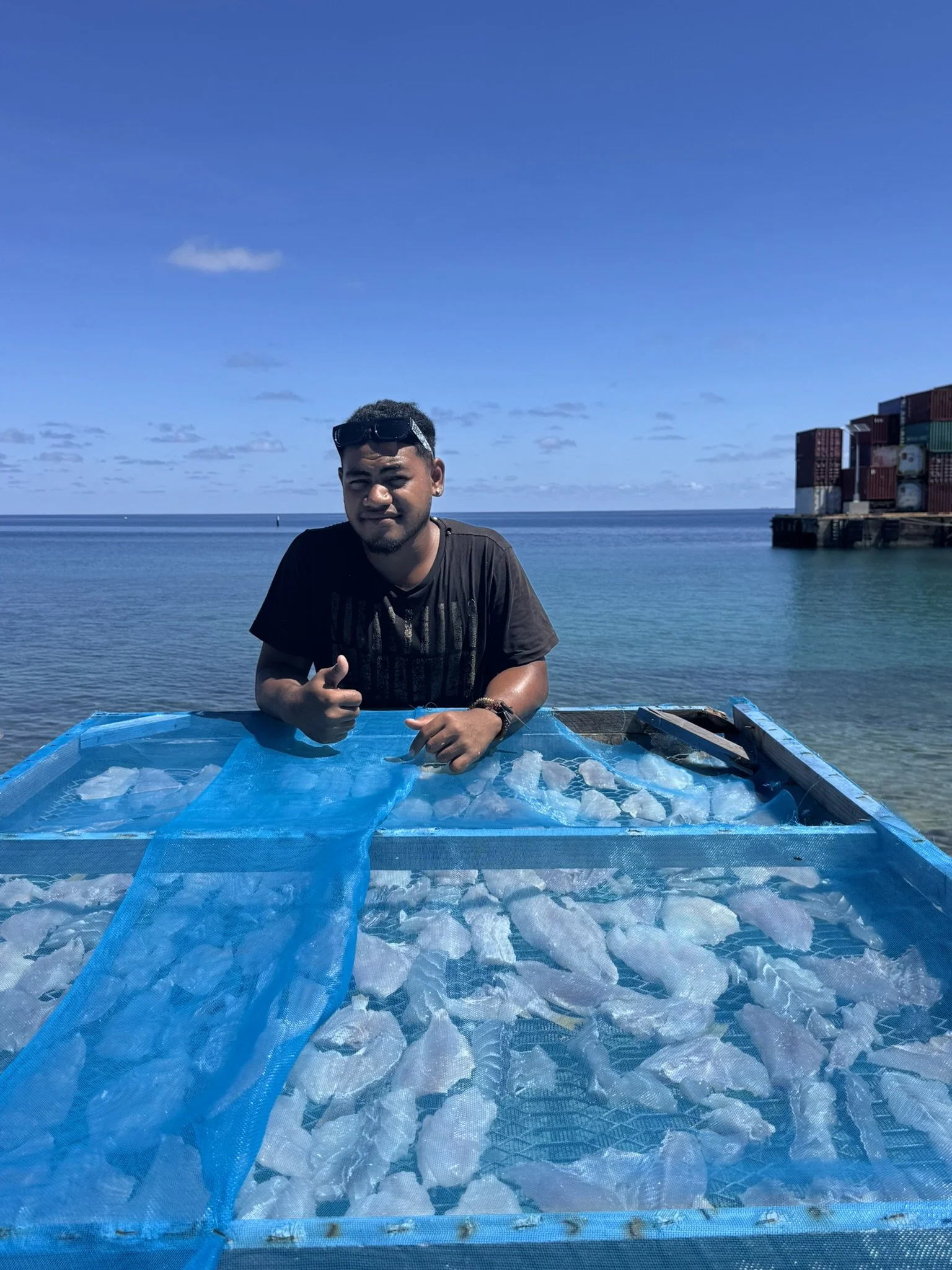 A man with sunglasses on his head gives a thumbs-up while standing behind a large blue container filled with ice, near a body of water with a shoreline and stacked shipping containers in the background.