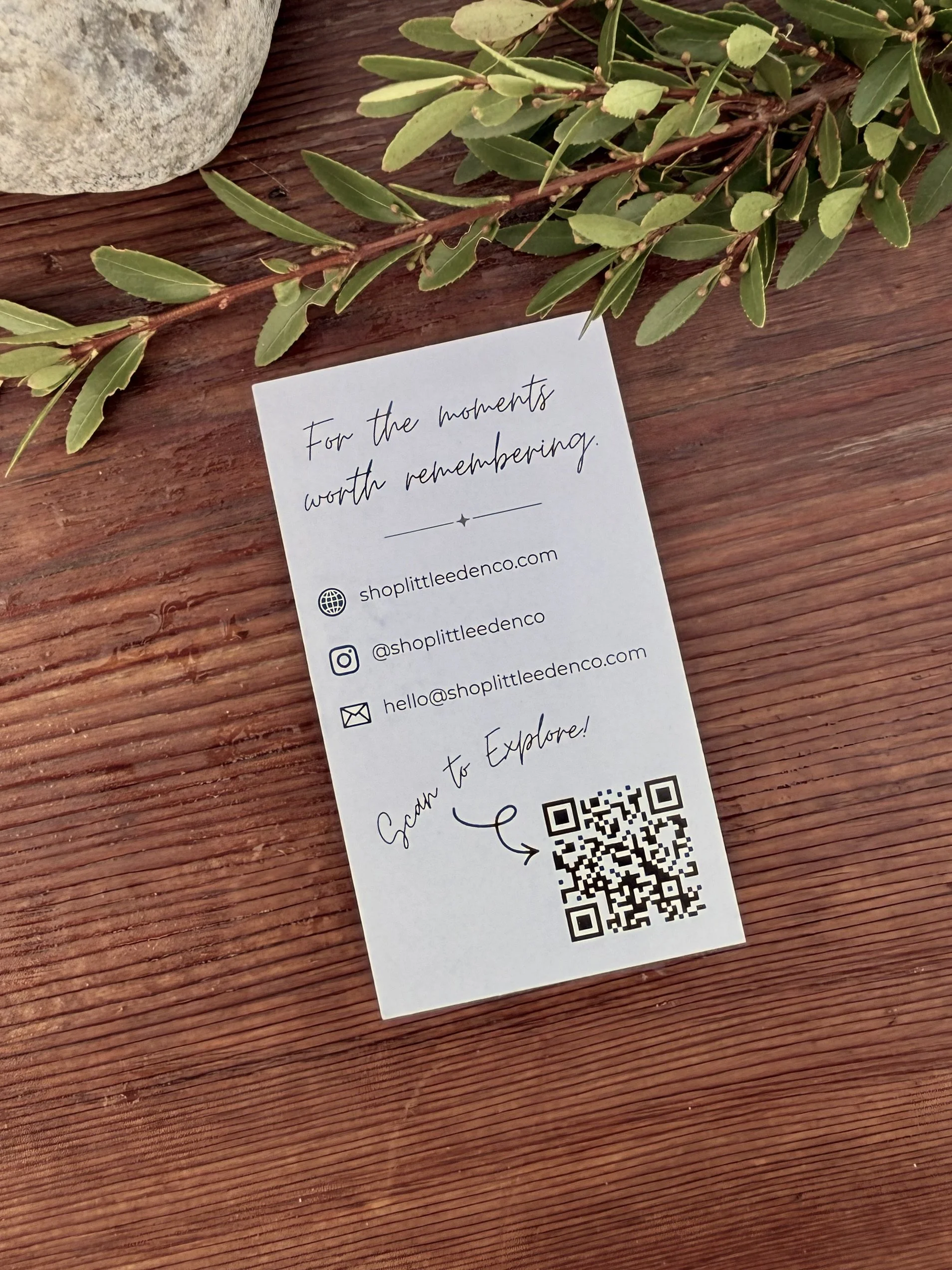 A gift tag with a handwritten message that reads 'For the moments worth remembering' on a wooden surface, with some green leaves and a stone nearby.