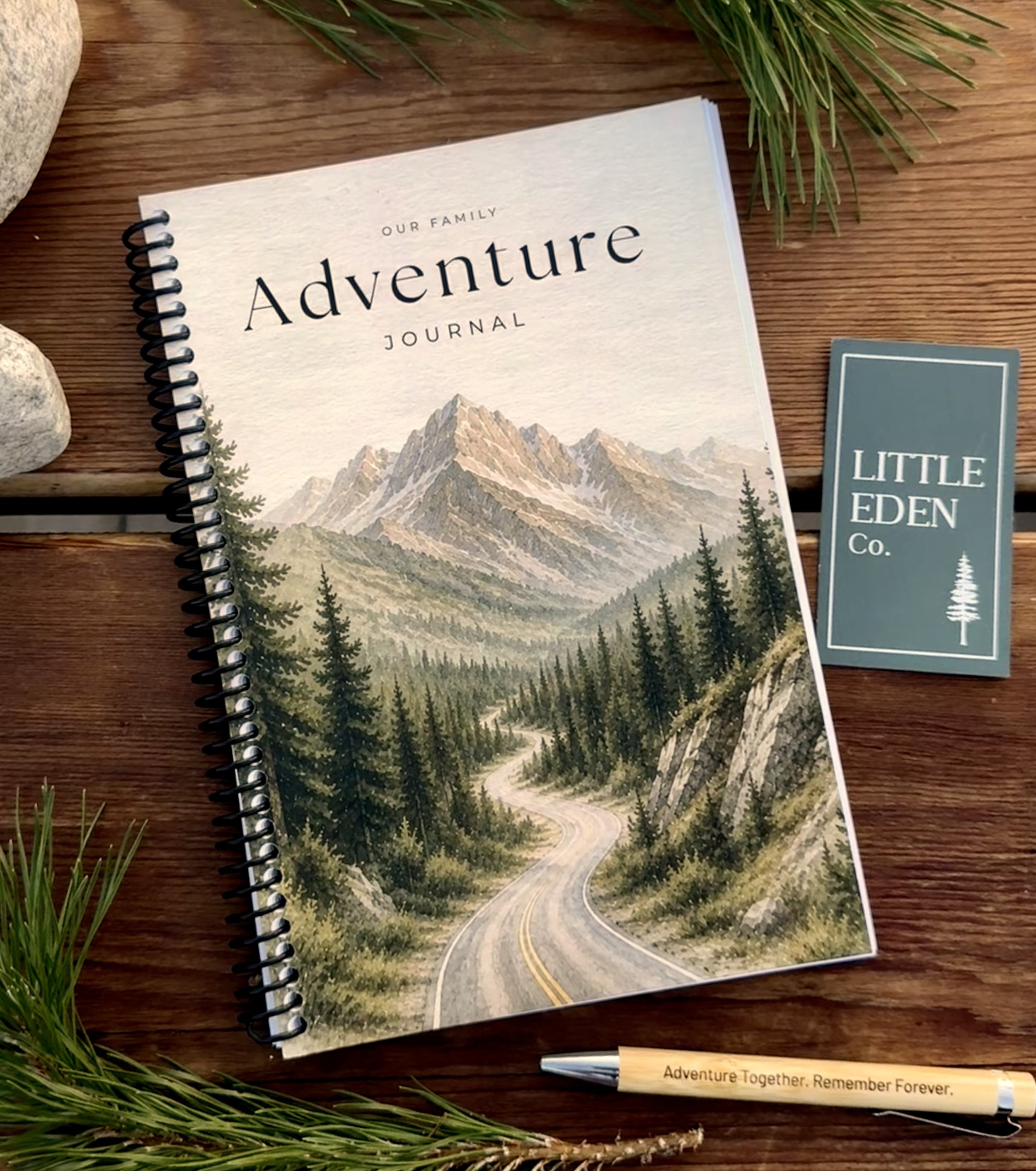 A spiral-bound journal titled 'Our Family Adventure Journal' on a wooden surface, with a mountain landscape cover, surrounded by small rocks, green grass, and a pen with the words 'Adventure Together. Remember Forever.'