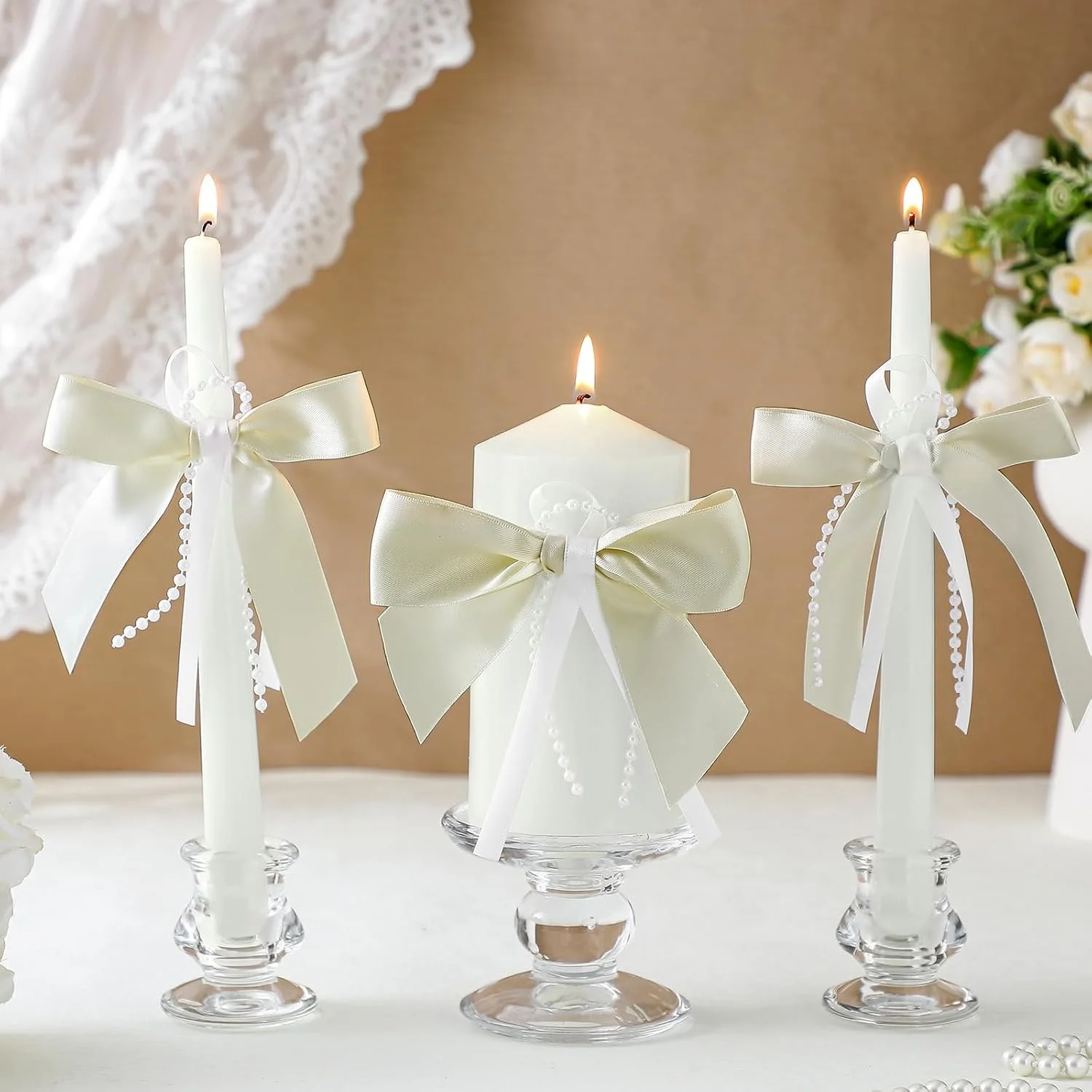 White candles decorated with cream-colored ribbons and pearl beads, arranged on glass holders, with a blurred floral arrangement in the background.
