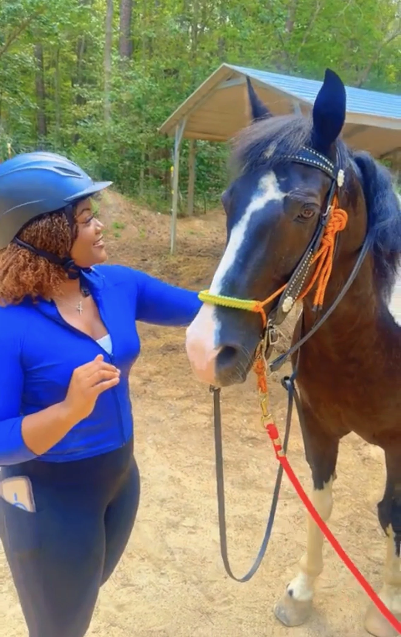 A woman wearing a blue shirt and a helmet is smiling and talking to a black and white horse. The horse has a riding bridle with decorative leather and orange rope, and its right front hoof is visible in the foreground. The scene takes place outdoors in a wooded area with trees and a small wooden shelter in the background.