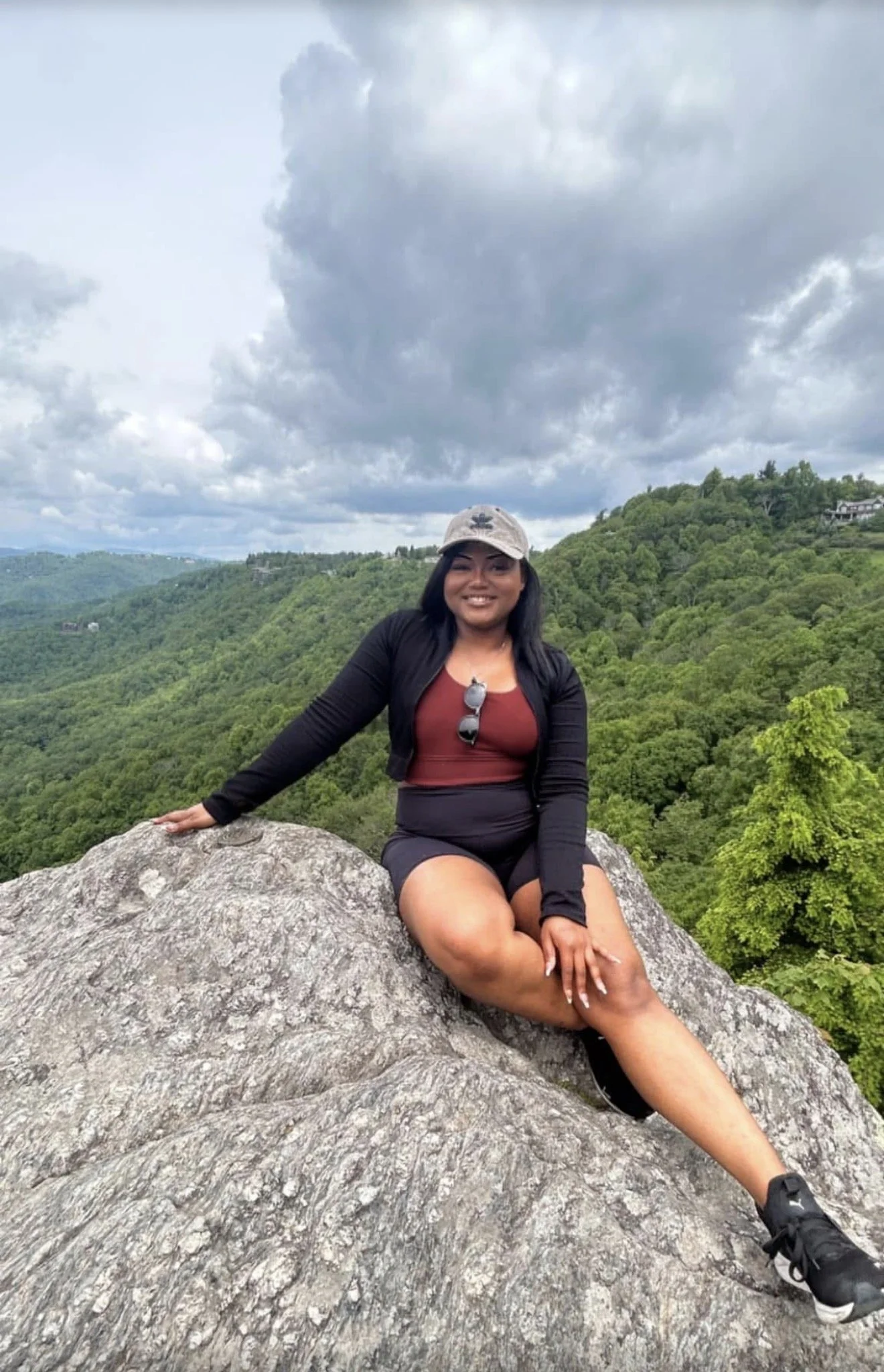 A woman sitting on a large rock in a forested mountainous area with cloudy sky above. She is smiling, wearing a gray cap, black jacket, burgundy top, black shorts, and black sneakers, with sunglasses hanging from her top.