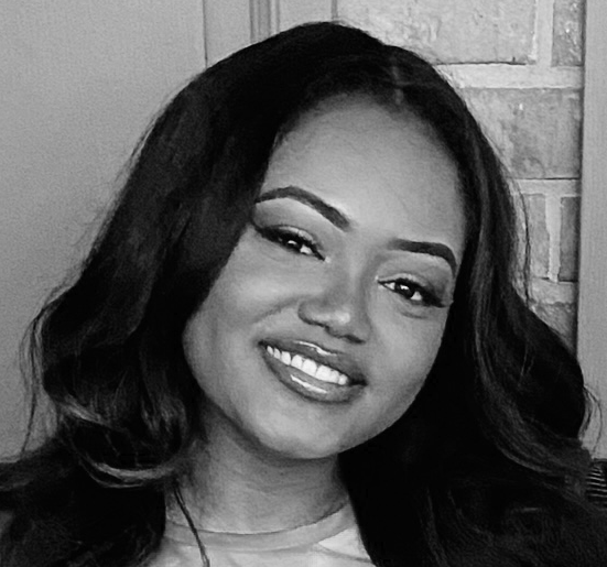 Black and white portrait of a smiling woman with long wavy hair, wearing a light-colored top, in front of a brick wall.