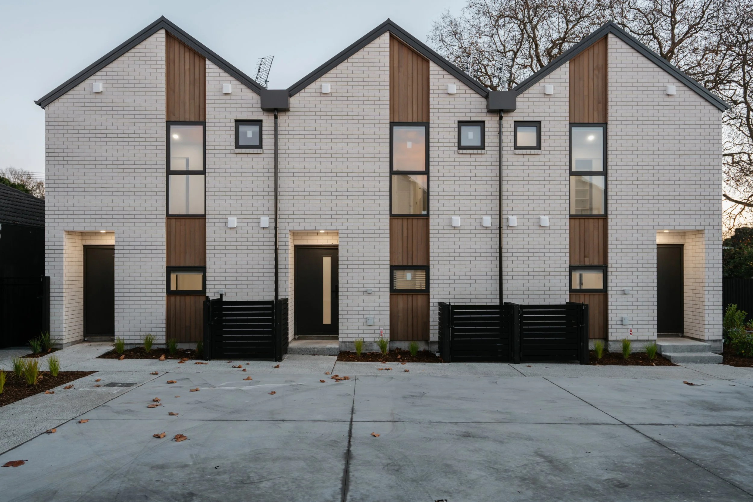 A modern multi-story residential building with a brick and wood exterior, large windows, and black gates in front, situated in a suburban neighborhood during late afternoon or early evening.