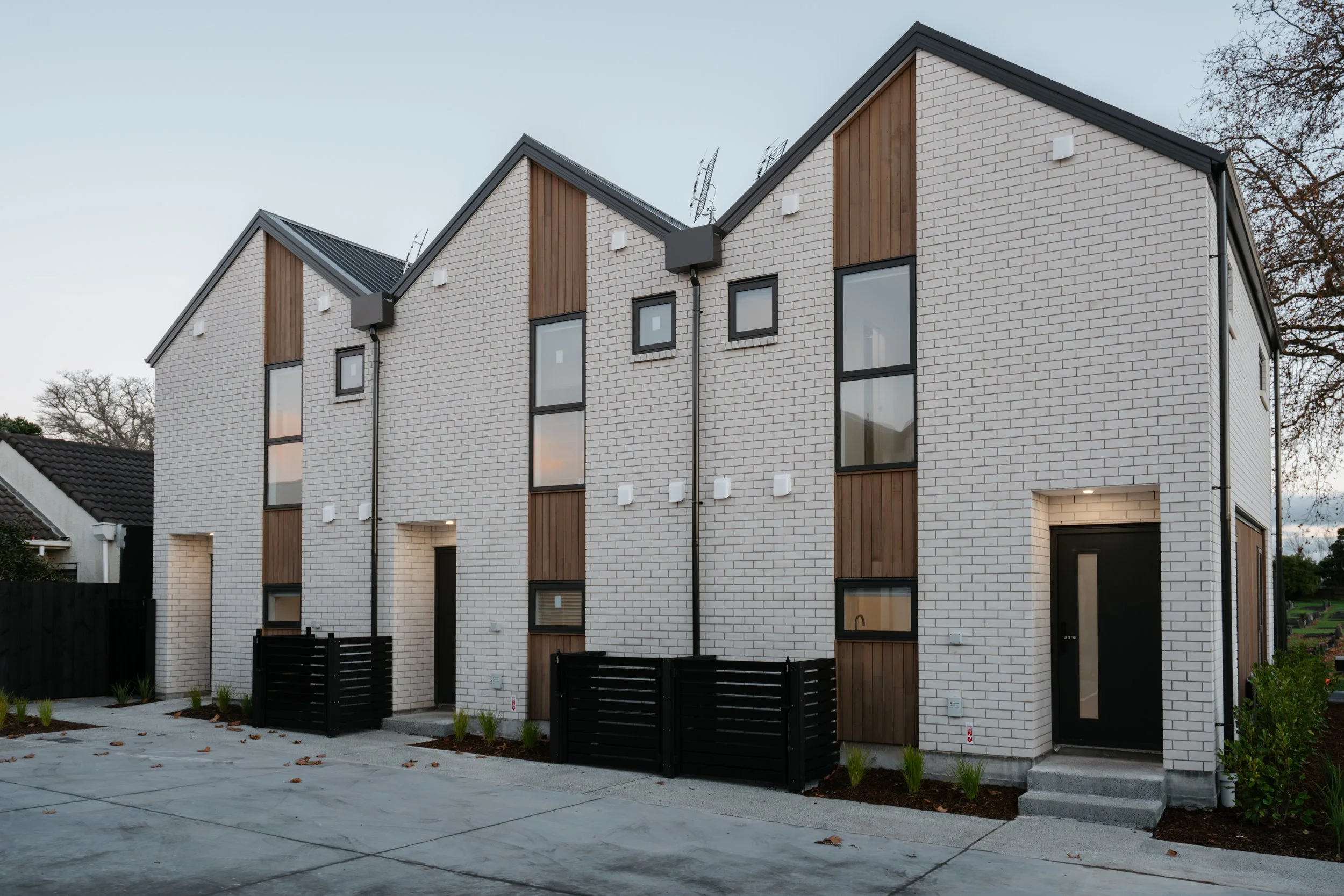 Modern multi-story residential building with white brick walls, black window and door frames, and wooden accents. Fenced yard with planting and paved patio area.