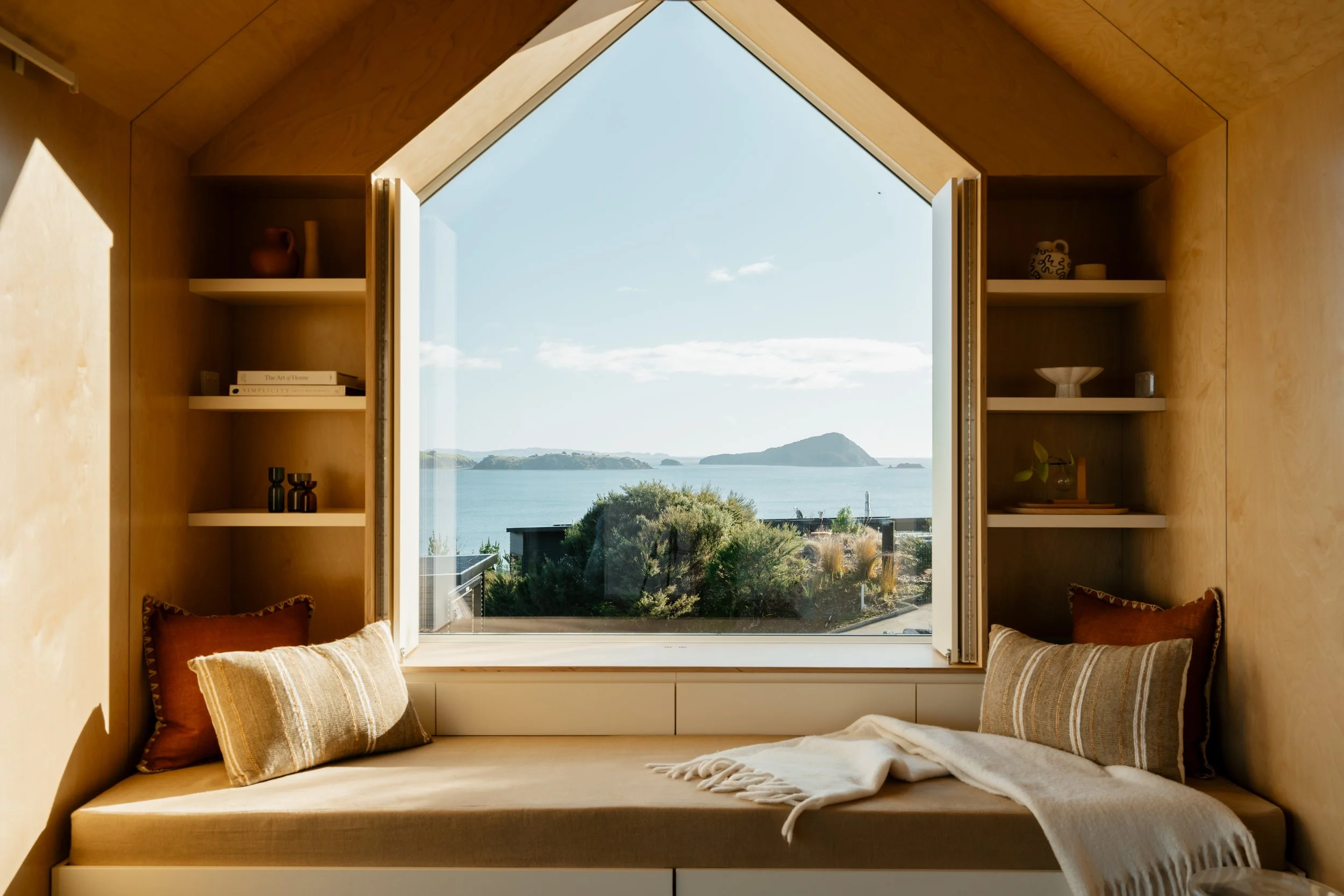Interior view of a cozy nook with beige cushions and pillows, built-in shelves, and a large window with a scenic ocean and island view, under a clear sky.