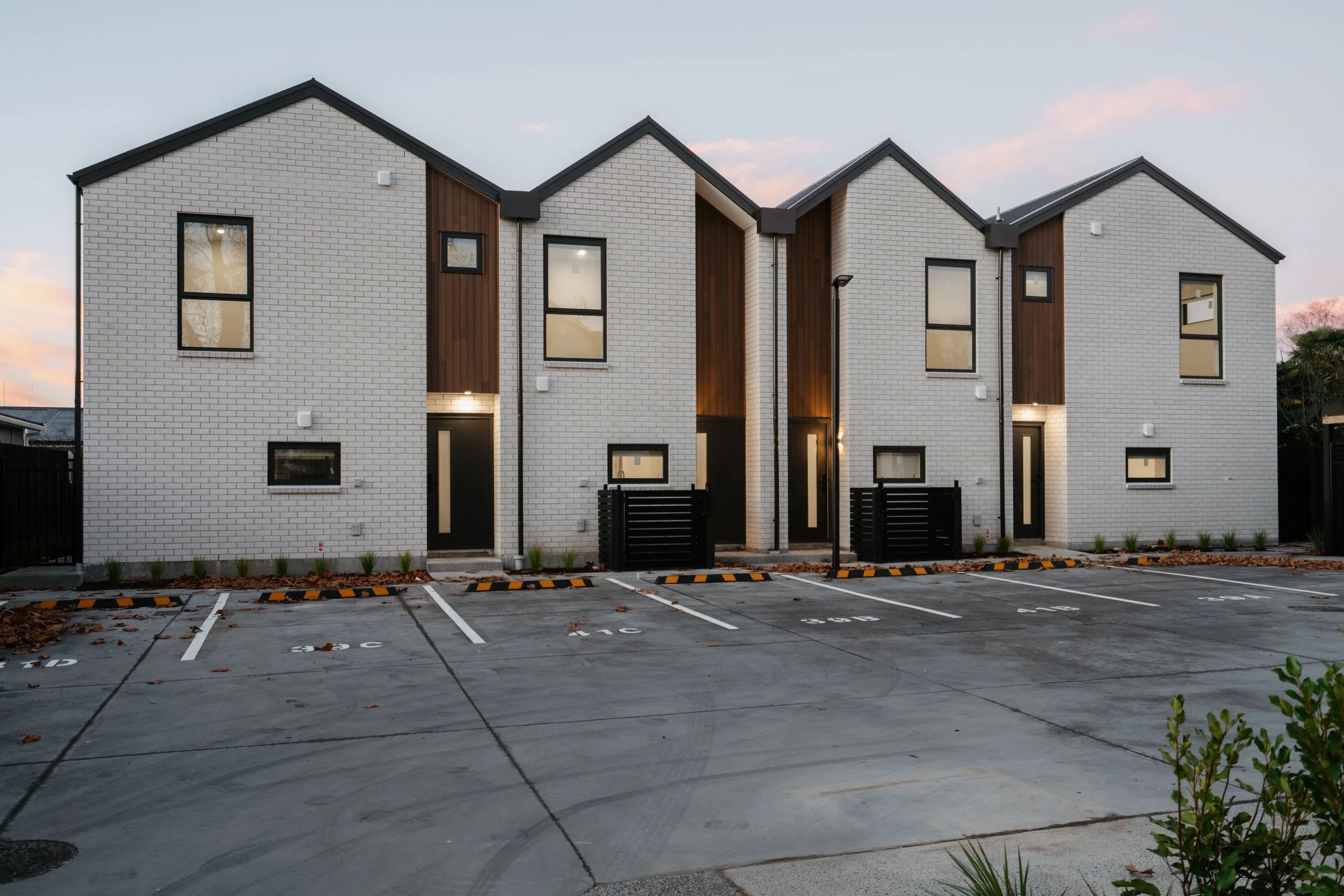 Modern white brick townhouses with black window frames and wooden accents, parking lot in front, evening sky.