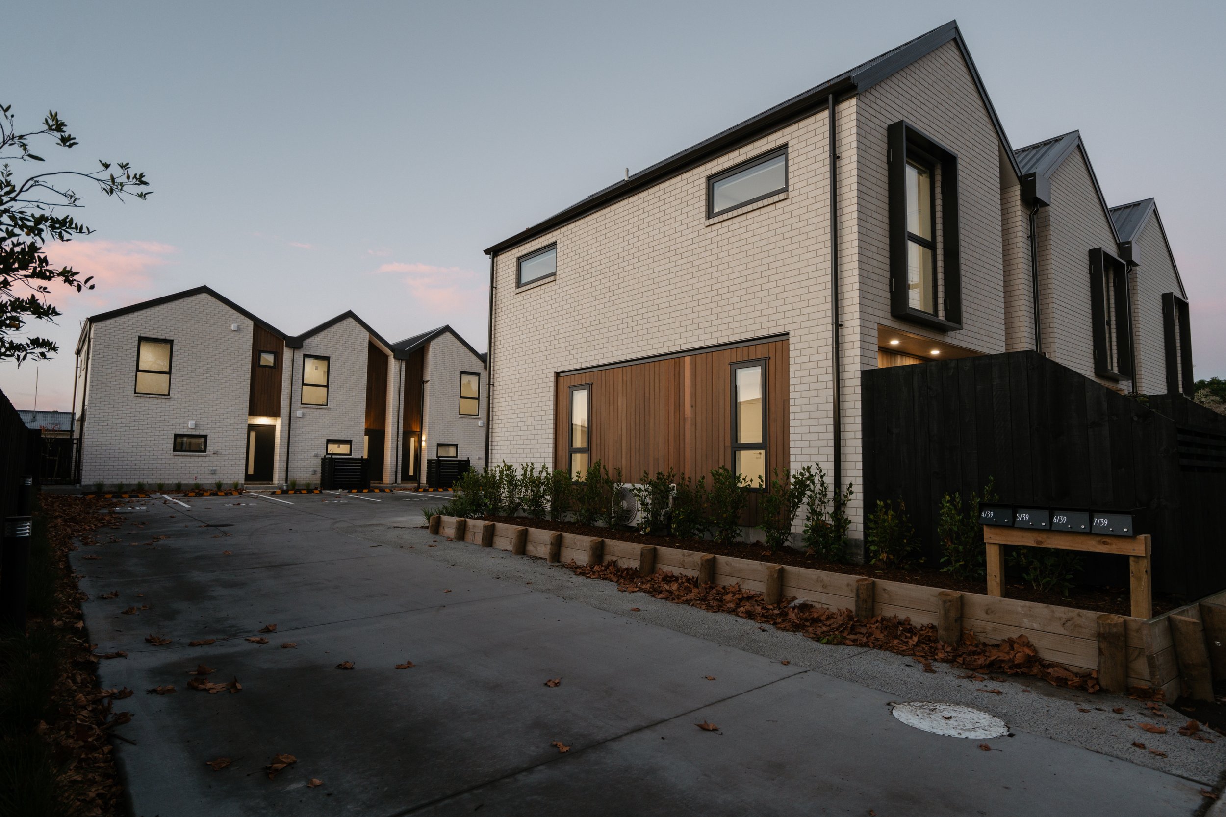 Modern townhouses with light-colored brick exteriors and dark-framed windows, located in a residential area during dusk. The street in front is paved, with some fallen leaves and small plants along the edges. There are mailboxes visible on the right 