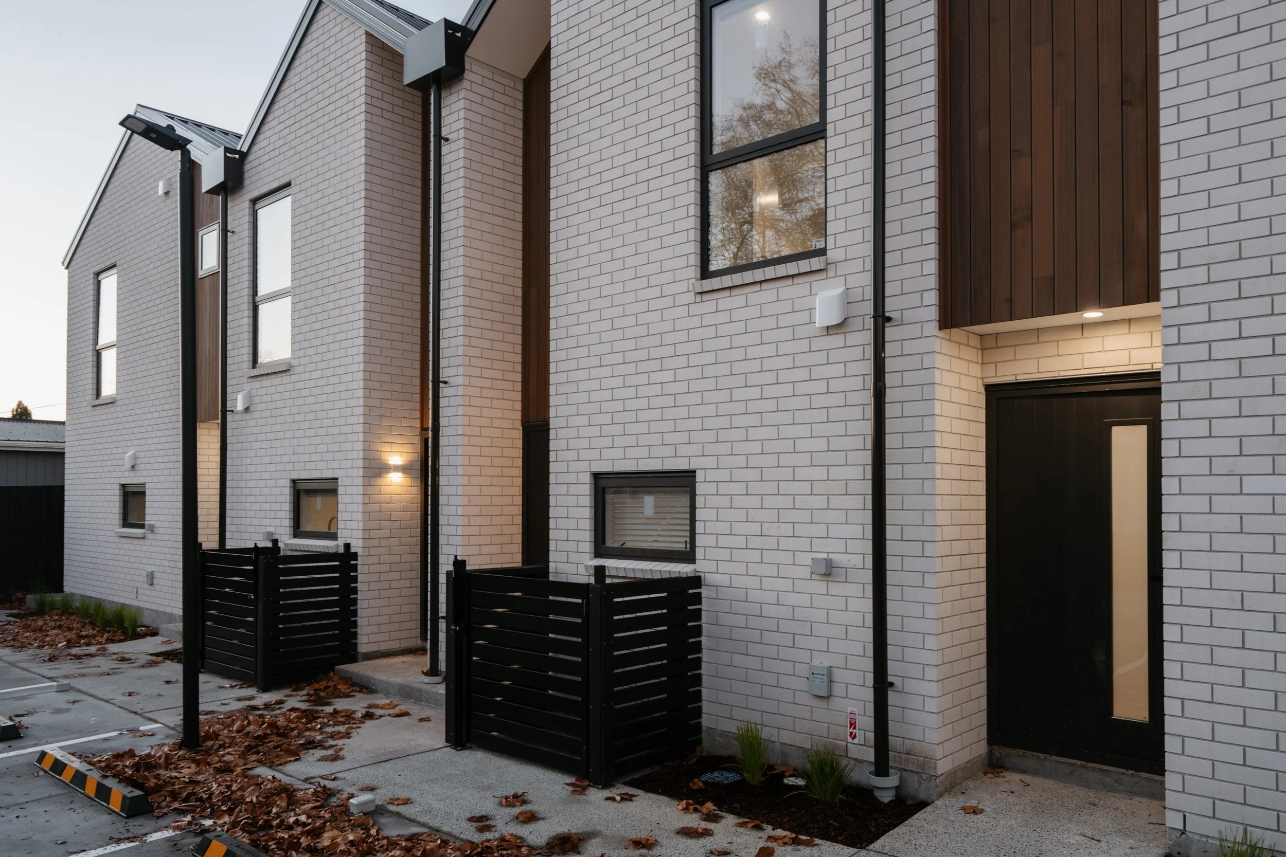 Exterior view of modern residential building with white brick walls, black-framed windows, and wooden accents. There are small fenced yards, parking spaces, and street lighting in the foreground. Fallen leaves are scattered on the ground.