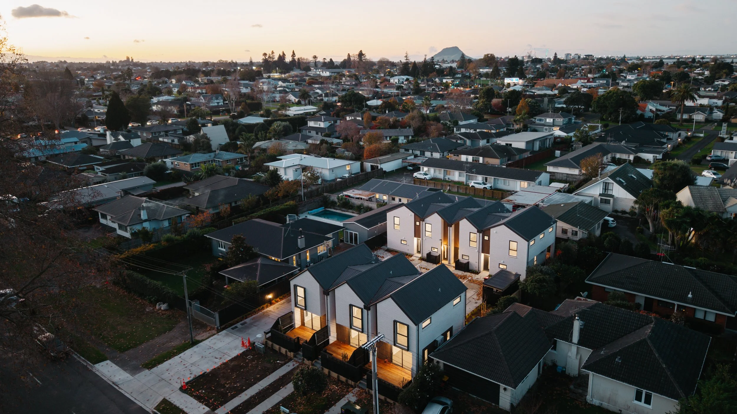 Aerial view of a residential neighborhood during dusk showing a mix of modern and traditional houses, with some lights on and a mountain in the background.