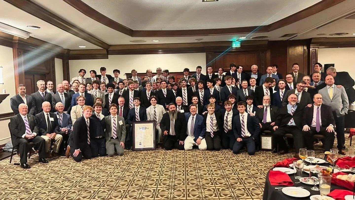 Group of men and boys dressed in suits and ties posing for a photo in a banquet hall, with tables set for an event in front.