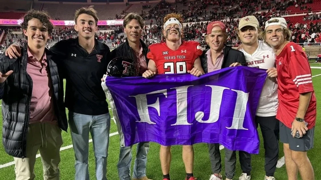 Group of young men standing on a football field, holding a purple FIJI fraternity flag, celebrating after a football game, with fans in the stands in the background.