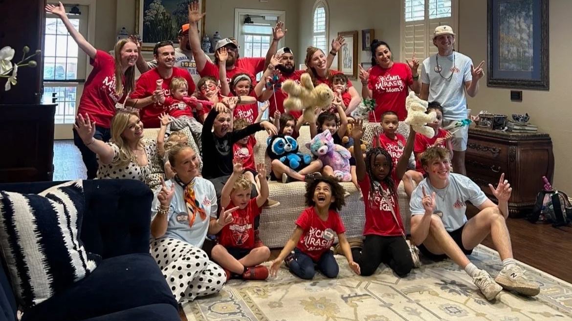 Group of children and adults posing happily indoors, with some holding plush toys and others making peace signs. Many wear red shirts that say "Camp Magic Mom" or similar. The atmosphere is cheerful, suggesting a camp or celebration event.