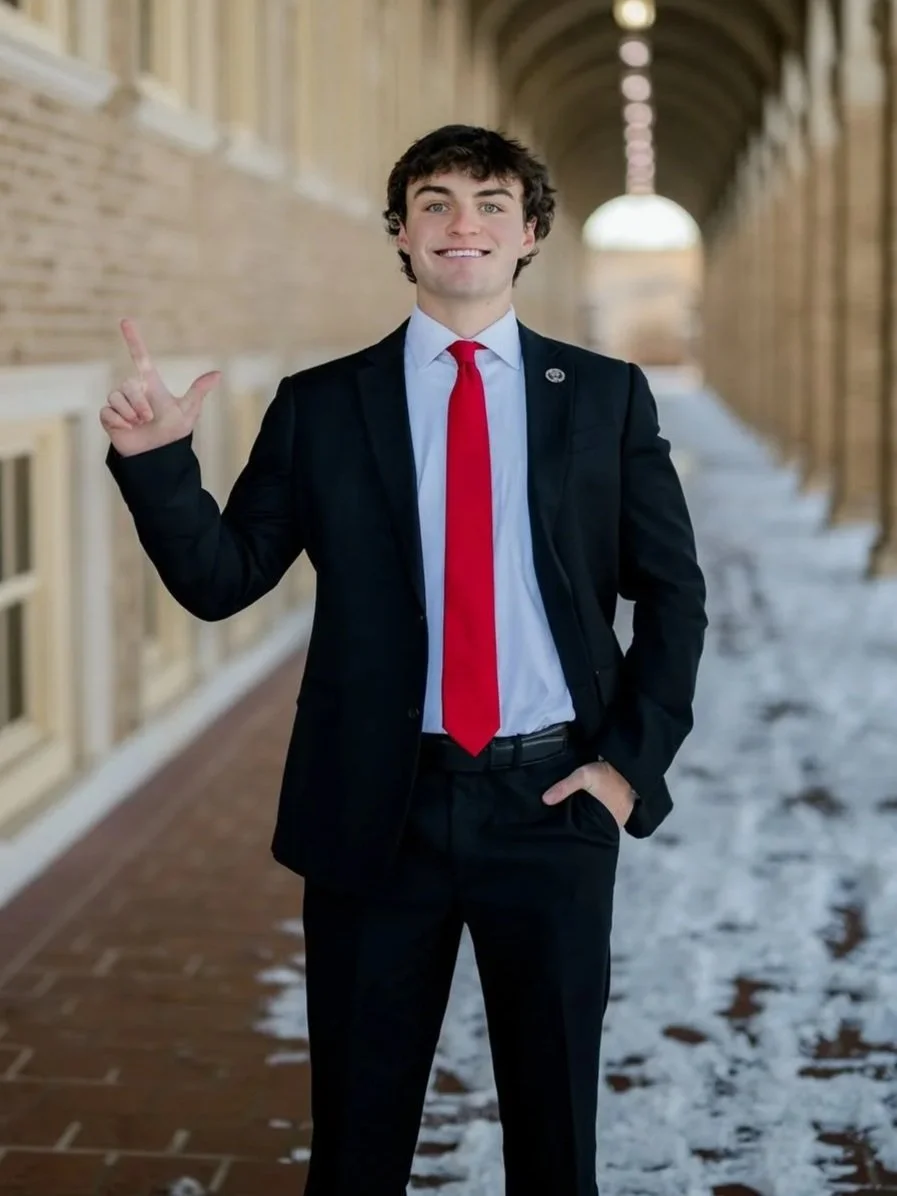 Young man in a black suit with a red tie standing outdoors in a covered walkway with brick walls, snow on the ground, smiling and making an L-shape with his left hand.