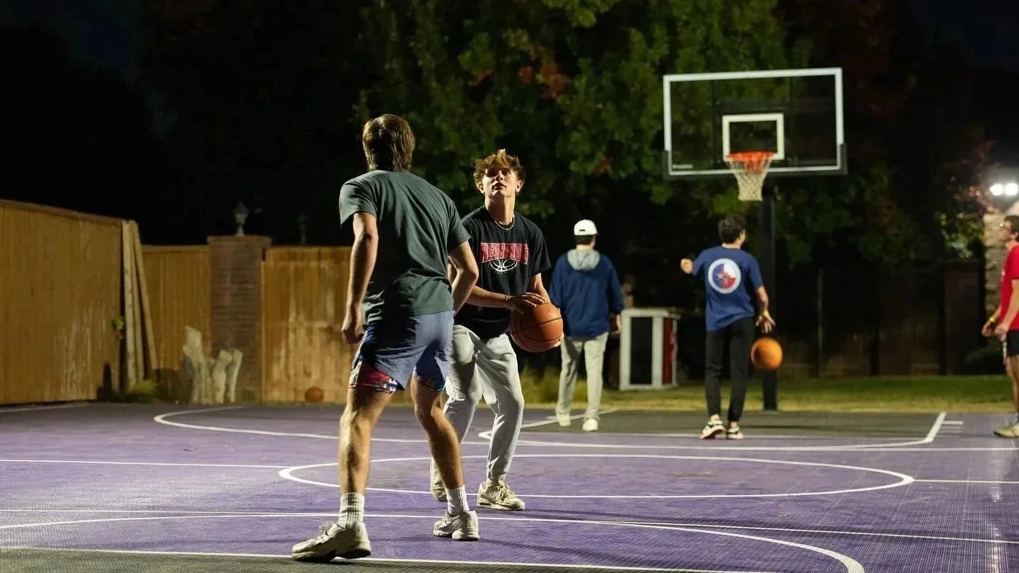 A group of young men playing basketball on an outdoor court at night. One player is holding a basketball, and a few others are standing nearby. The court is surrounded by a wooden fence, and there are trees in the background.