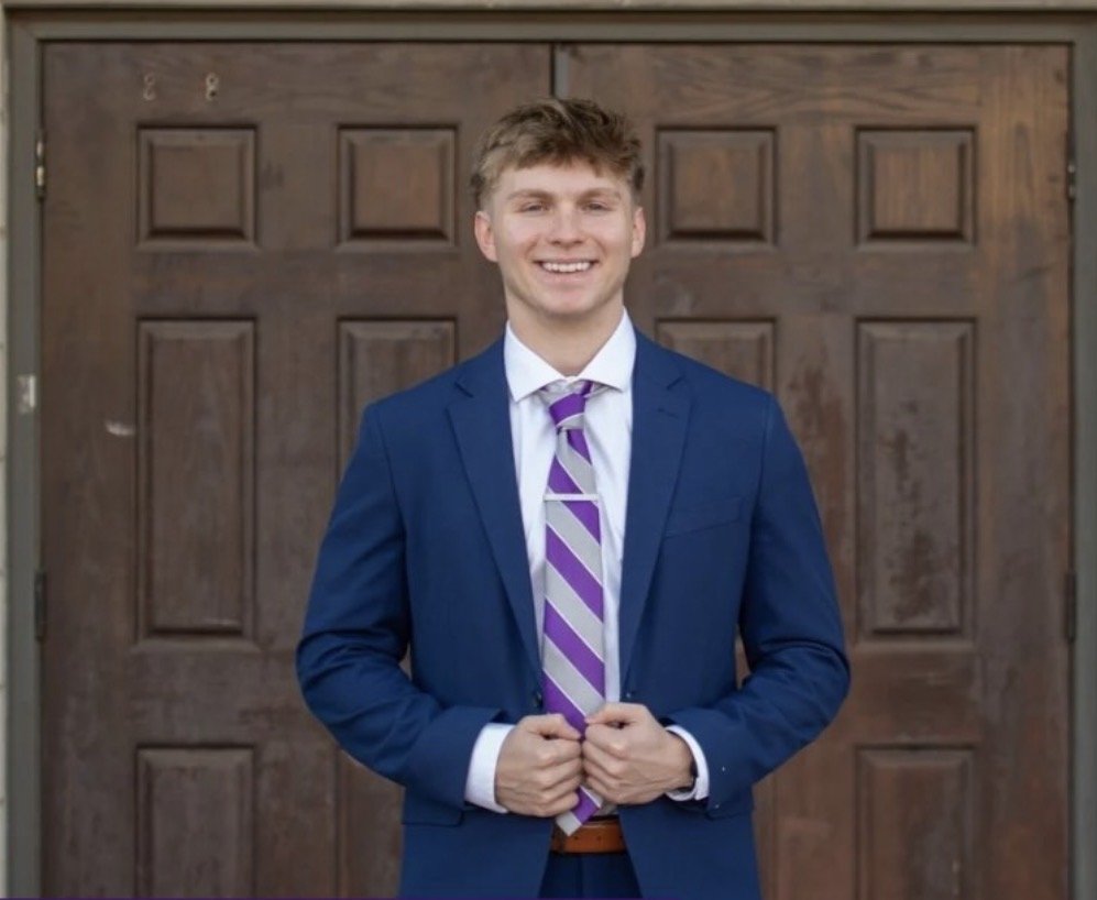 A young man in a blue suit, white shirt, and purple-striped tie standing in front of a wooden garage door, smiling and holding his jacket lapels with both hands.