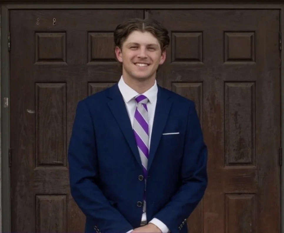 A young man in a dark blue suit and purple striped tie standing in front of a wooden door, smiling at the camera.