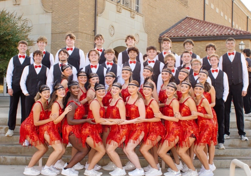 Group of young men and women dressed in vintage-style outfits, posing outside on steps in front of a historic building. The women are wearing red fringed dresses with headbands, and the men are wearing black vests, white shirts, red bow ties, and black trousers.