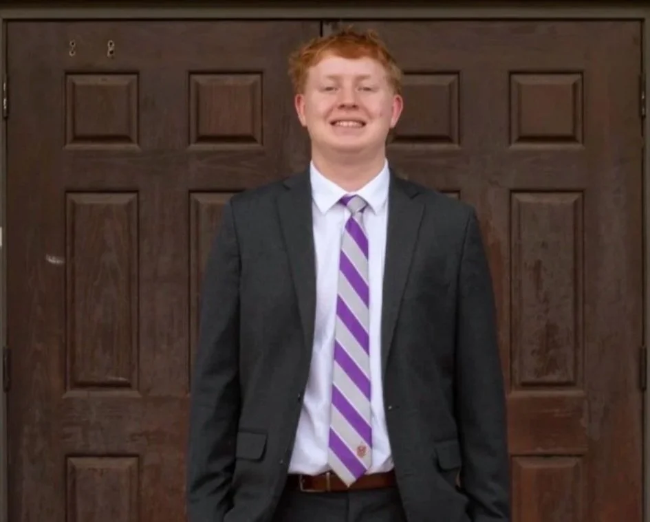 Young man with red hair smiling, dressed in a dark suit with a white shirt and a purple striped tie, standing in front of a brown wooden door.