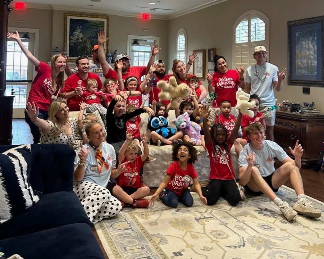 Group of children and adults gathered in a living room, celebrating with stuffed animals and raising their hands, some wearing red shirts, in a joyful, festive atmosphere.
