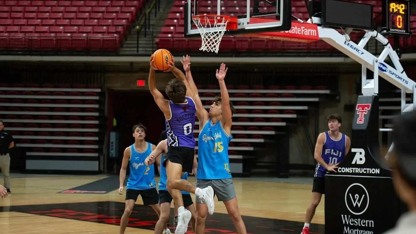 A basketball game with players jumping for a rebound or shot near the basket. One player in purple is holding the ball, while a player in blue attempts to block or defend. Other players and a coach are visible in the background on a basketball court.