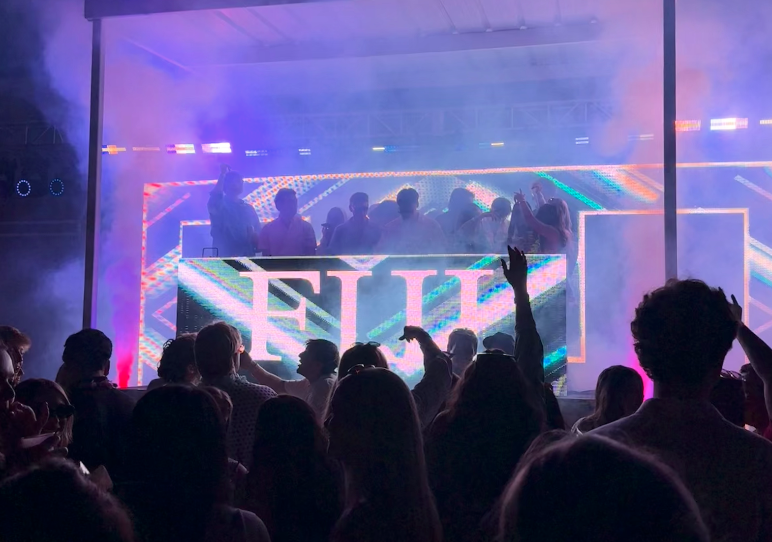A DJ performs on a stage with a large moving digital display at a lively music event, with a crowd dancing and raising their hands in front of the stage. Colorful lights and smoke create an energetic atmosphere.