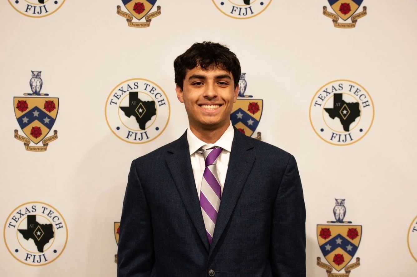 Young man in a dark suit with a white shirt and purple striped tie standing in front of a backdrop with seals of Texas Tech University in Fiji and shields with an owl and roses.
