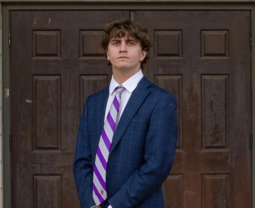 A young man in a blue suit with a white shirt and purple striped tie standing in front of a wooden door.