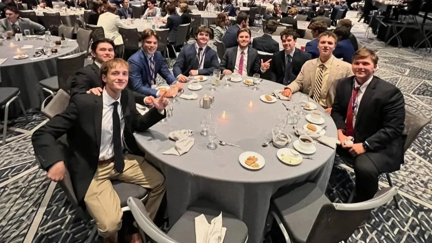 Group of ten young men in suits sitting around a large round table at a formal event.
