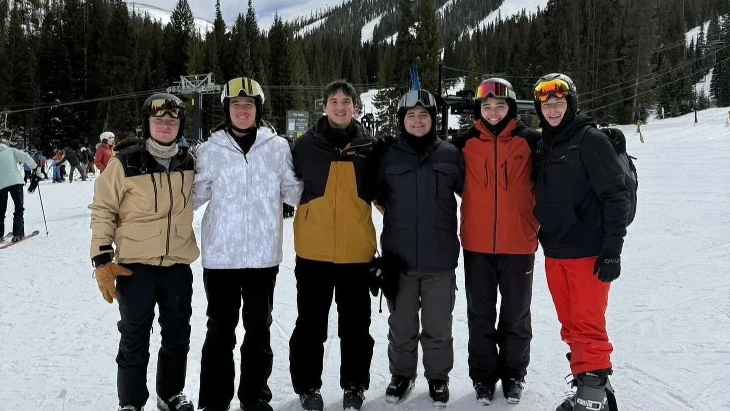 Six young men in winter clothing and ski gear standing on snow, with a snowy mountainside and pine trees in the background.