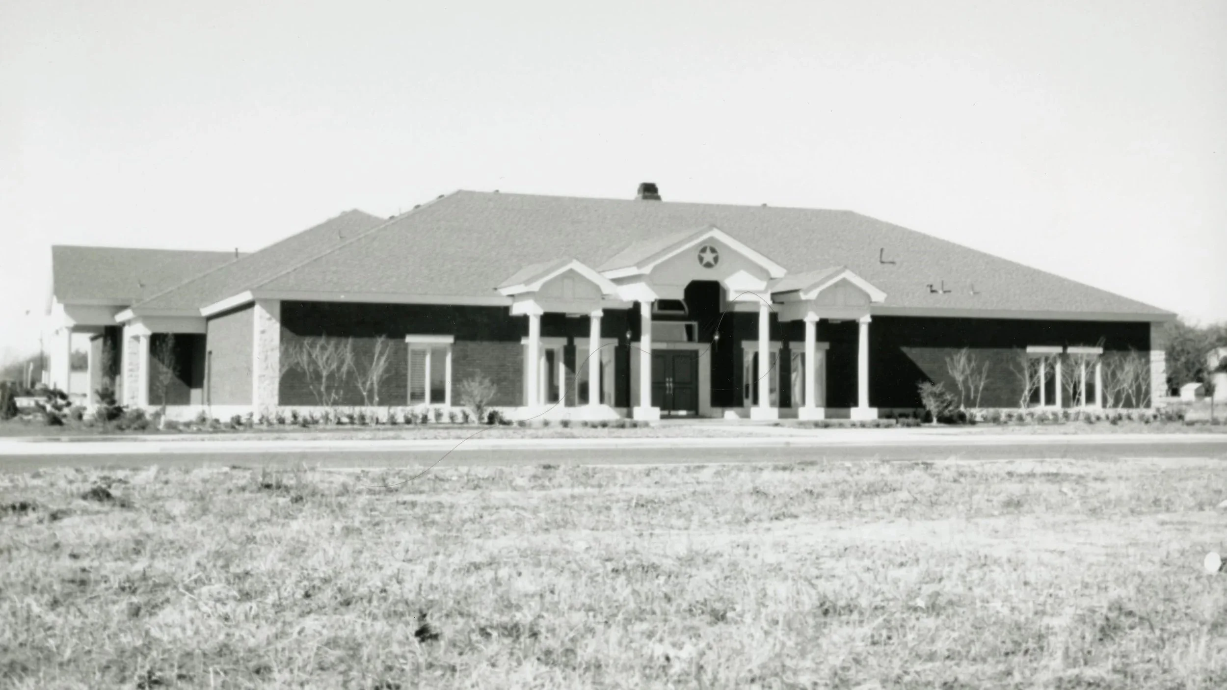 A large, single-story house with a symmetrical facade, front porch supported by multiple columns, gabled roof, and a star emblem above the entrance, set against a clear sky with a grassy lawn in the foreground.
