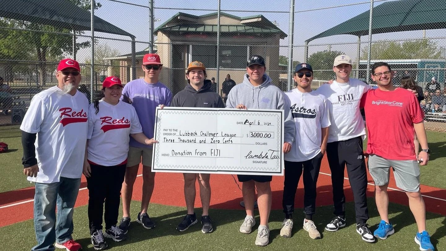 Group of nine people standing on a baseball field, holding a large donation check for $3,000 to Lubbock Challenger League, with a fence and a building in the background.