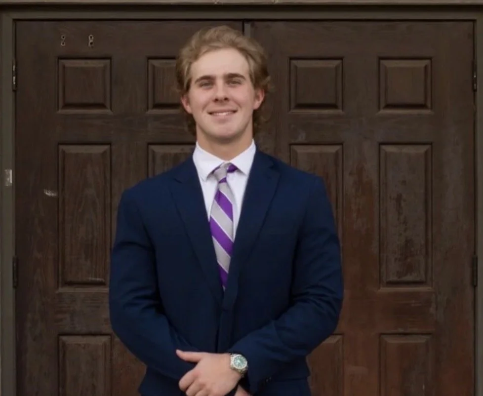 A young man in a navy suit with a purple and white striped tie, standing in front of a wooden door, smiling with arms crossed.