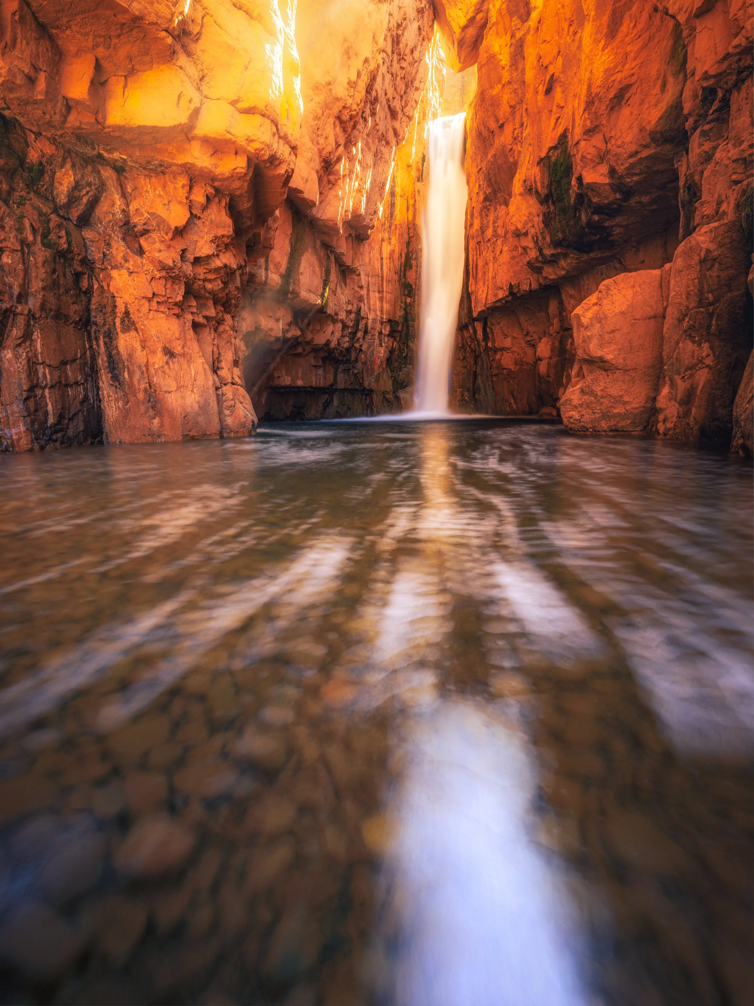 A tall waterfall cascading down a red rocky canyon with sunlight illuminating the rocks and reflecting on the water at the base.