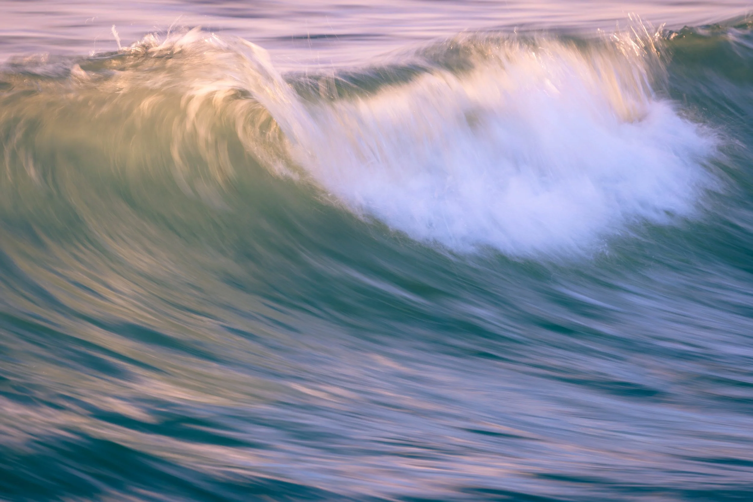 A close-up view of ocean waves during sunset or sunrise, with soft pink and purple hues in the sky reflected on the water.