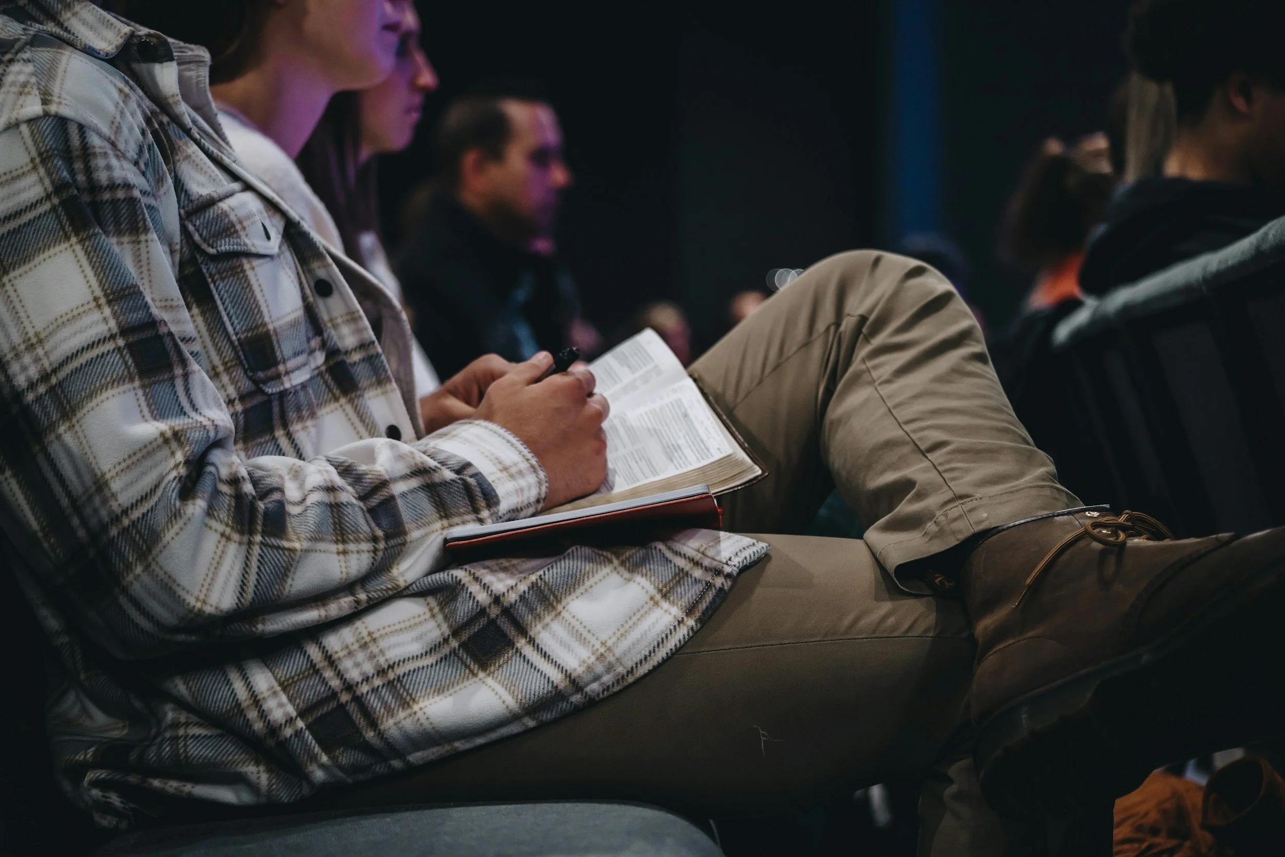 People sitting in a dark room, focusing on their notes or devices, possibly attending a presentation or lecture.