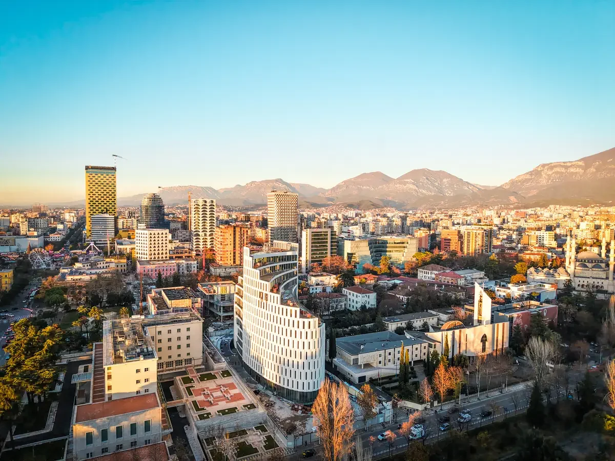 Aerial view of a city skyline with tall modern buildings, hills in the background, and a clear blue sky during sunset.