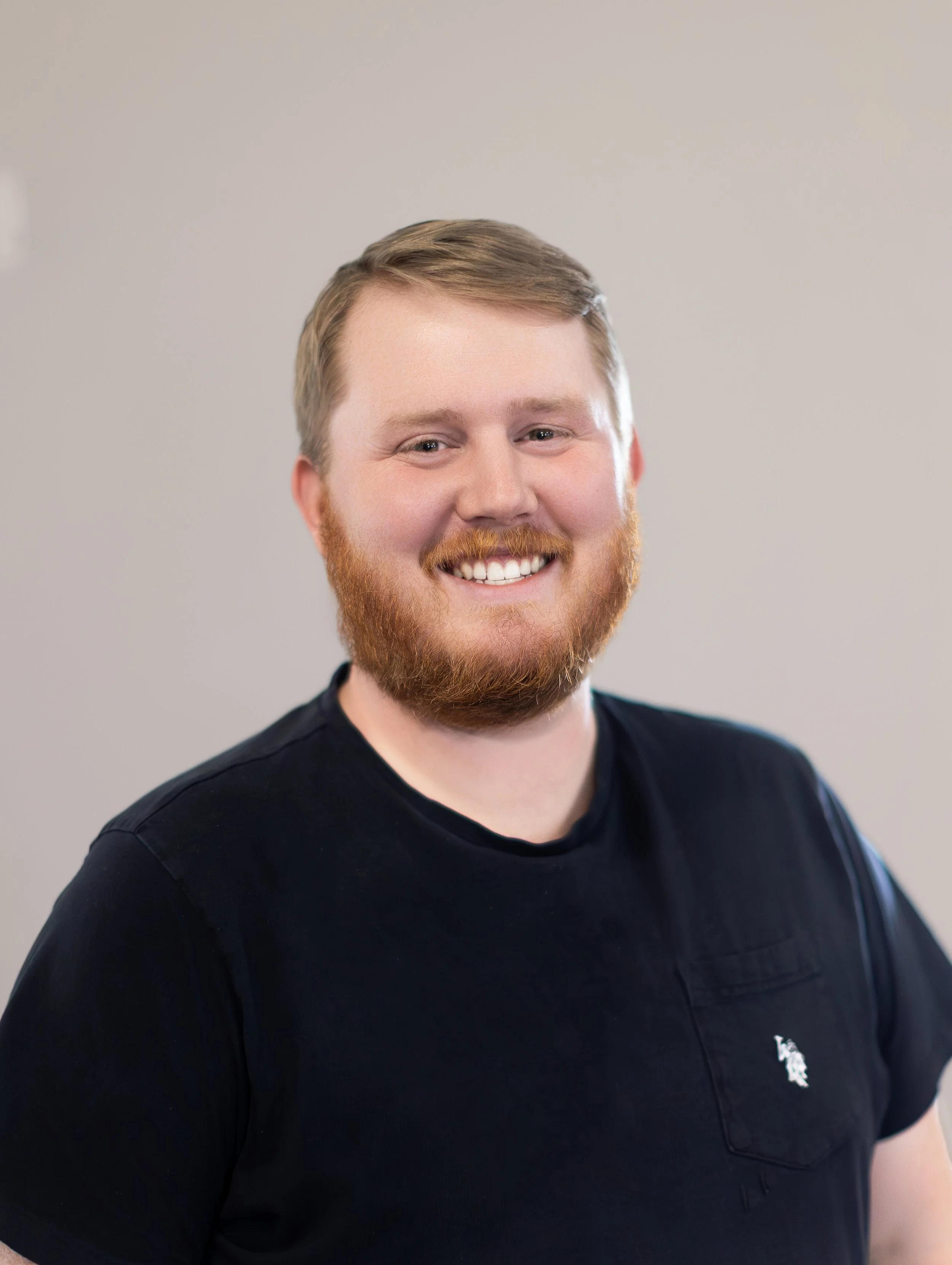 Close-up of a smiling man with red hair and a beard, wearing a black shirt with a white logo on the chest, against a plain light gray background.
