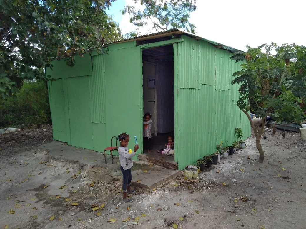 Three young children inside and outside a small house made of green corrugated metal panels, with trees and some cars visible around, and a dirt ground.