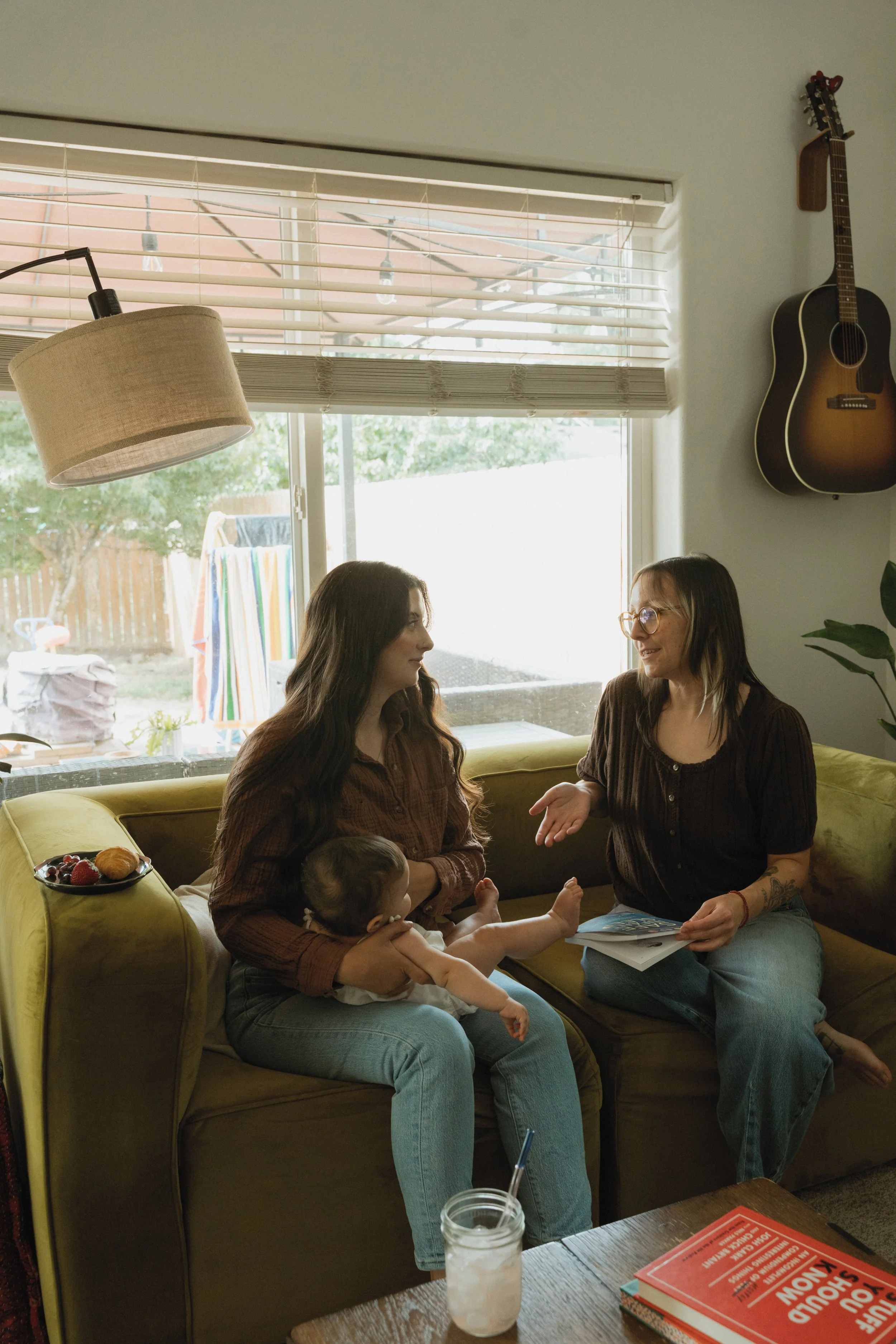 Two women sitting on a yellow couch in a living room, engaging in conversation. A small child is sitting on one woman's lap. There is a plate of berries and a pastry on the armrest of the couch. A glass jar with a drink and straw is on the table in front. A window with blinds lets in natural light, and a guitar hangs on the wall. Outside, a backyard with a clothesline and colorful towels is visible.