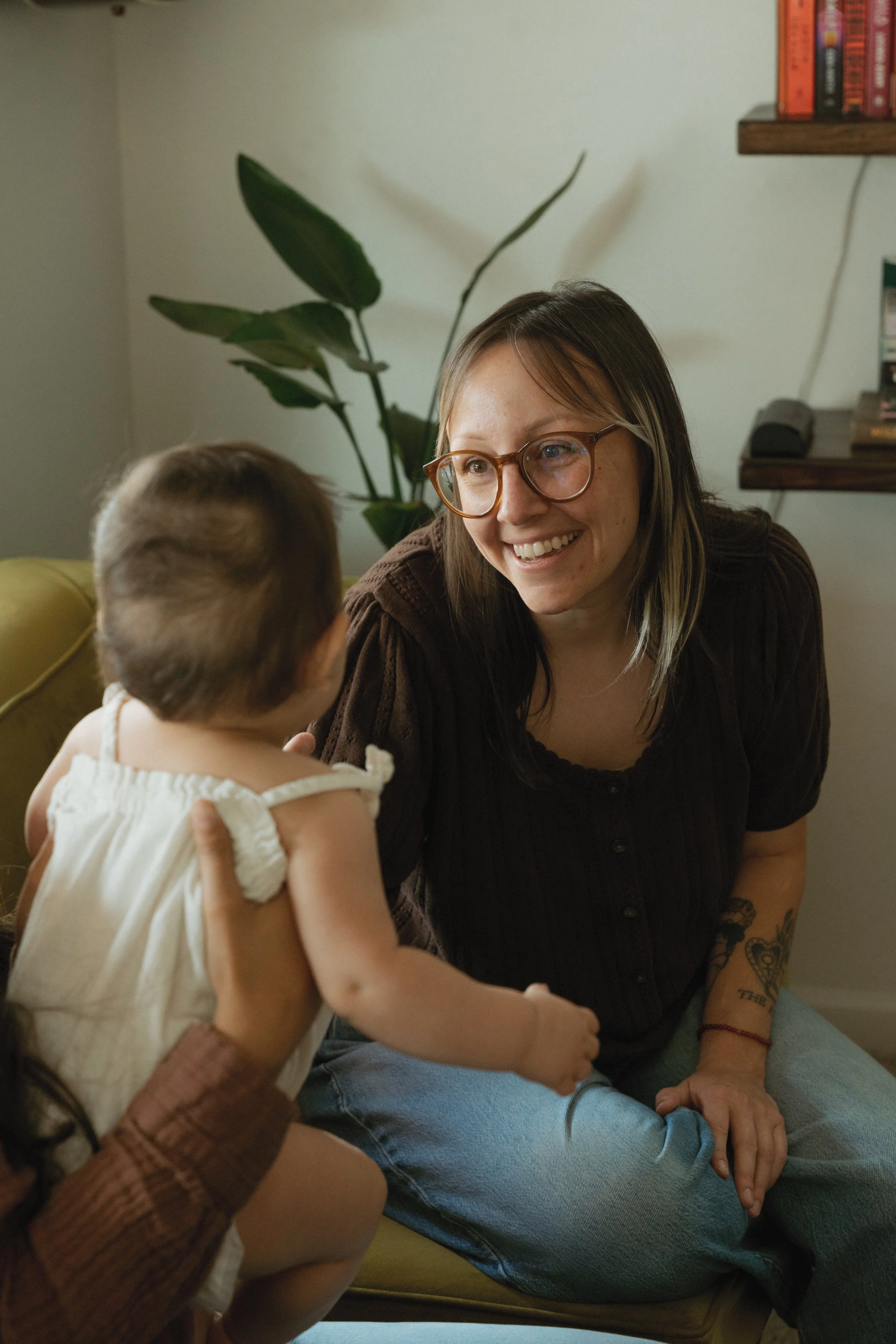 A woman with glasses smiling and engaging in conversation with a small child, who is being held by someone with darker skin and wearing a sleeveless white dress. They are seated on a yellow couch in a living room with a plant in the background and shelves with books.