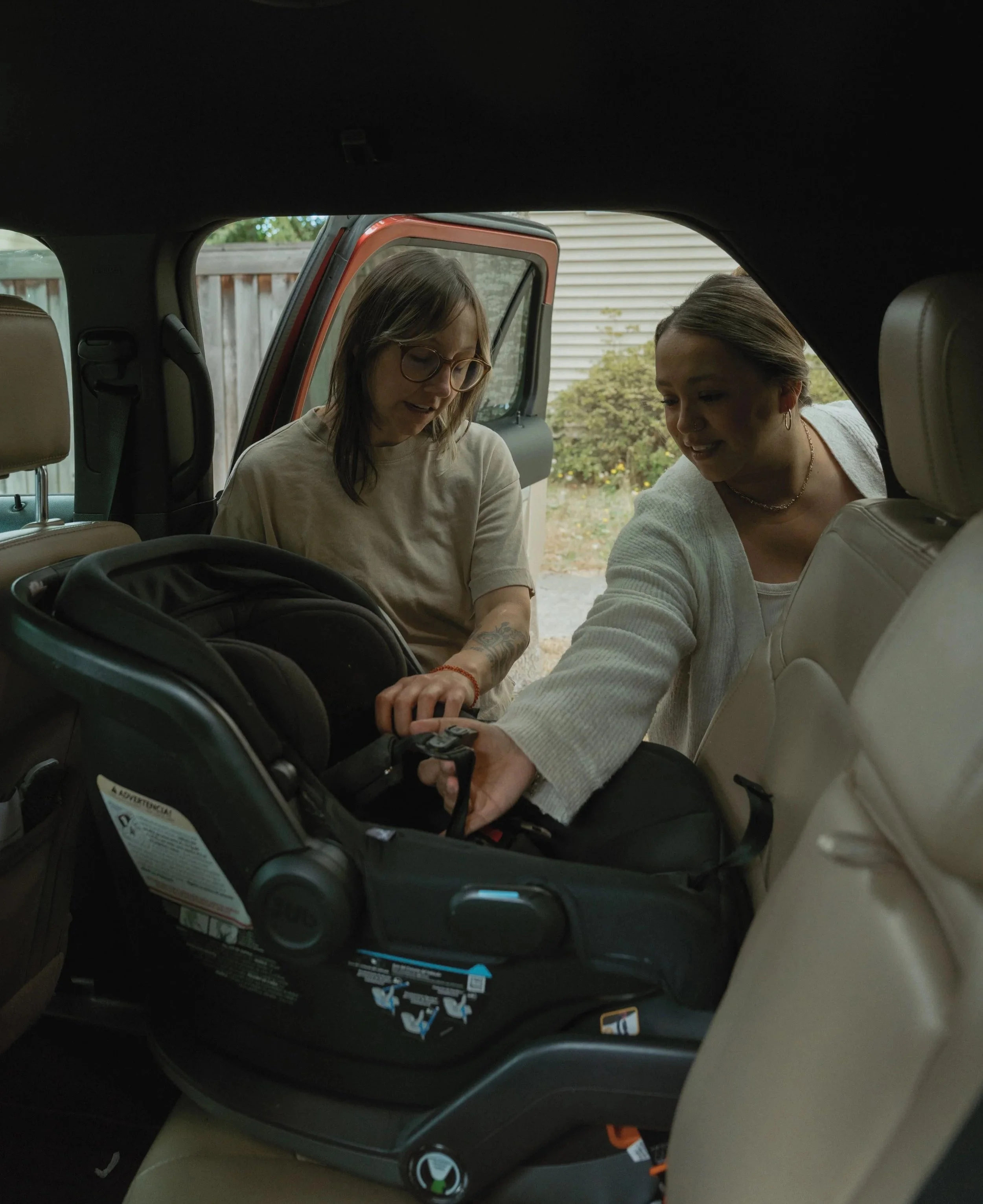 Two women standing outside a vehicle, one holding a car key near a child's car seat, with a wooden fence and house siding in the background.